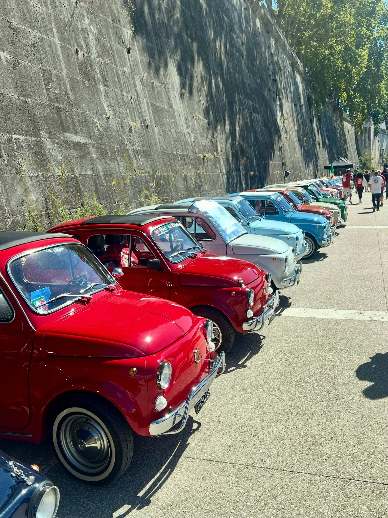 Vecchie auto fiammanti parcheggiate lungo un muro di pietra, con alcuni persone che passeggiano in background