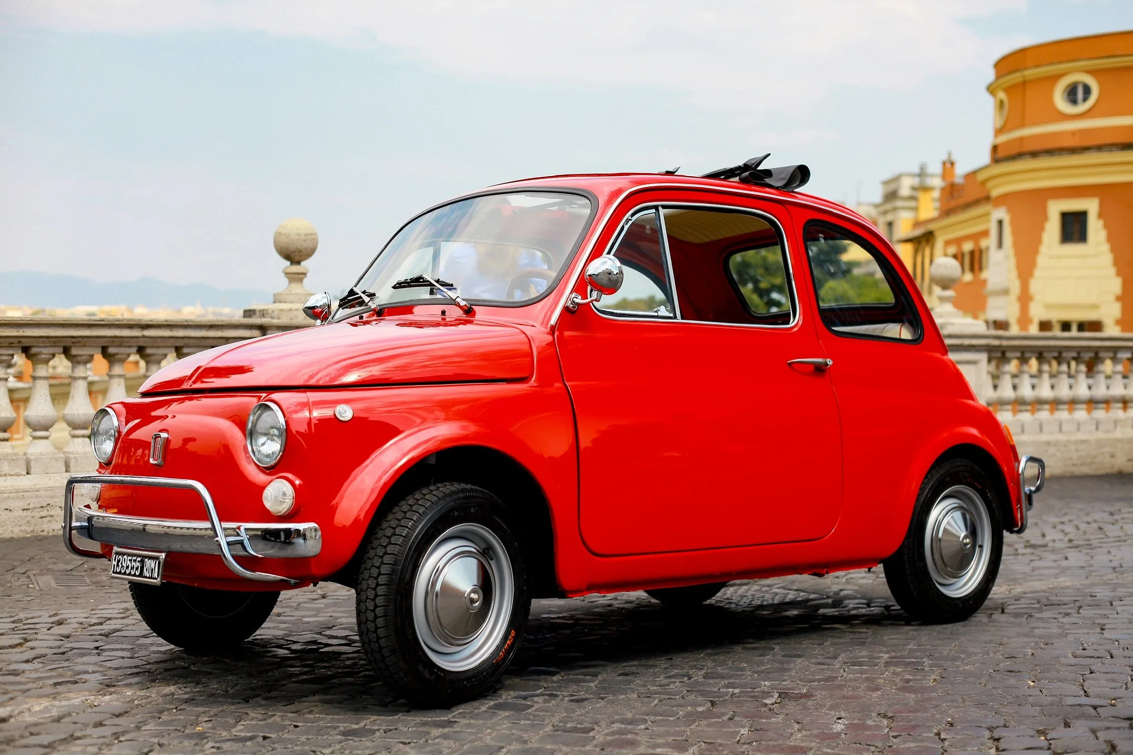 Auto rossa vintage Fiat 500 parcheggiata su strada di ciottoli con edificio giallo-arancione sullo sfondo.