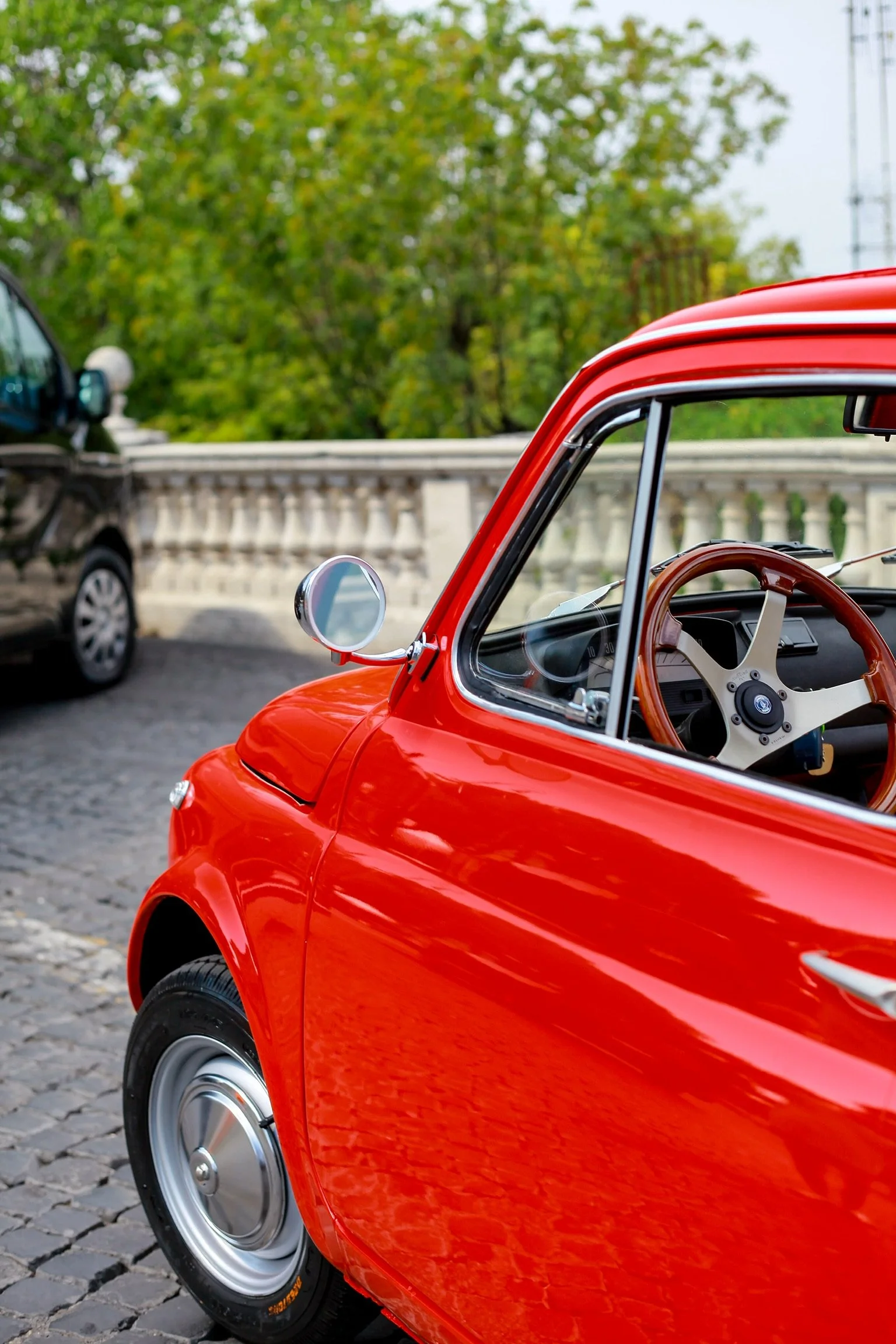 Auto rossa d'epoca con volante in legno parcheggiata su strada con ciottolato, con sfondo alberi verdi e un muretto bianco.