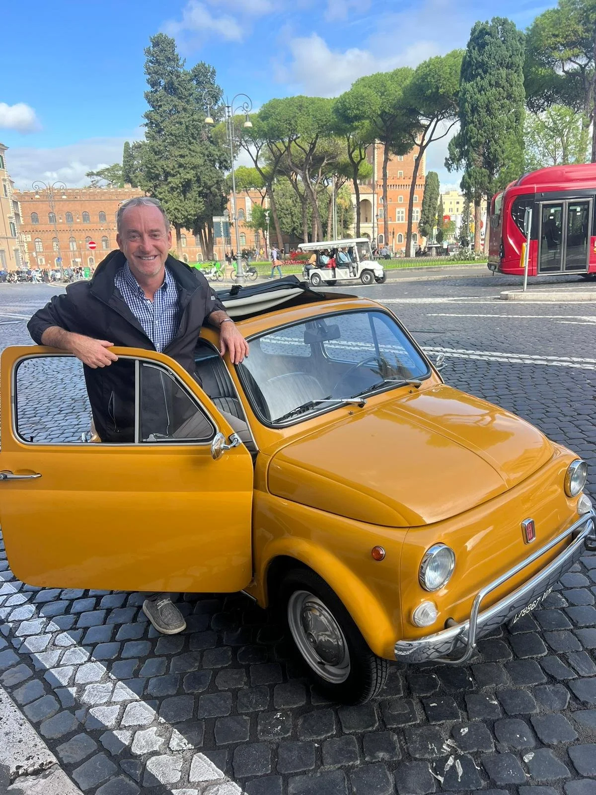 Uomo sorridente che si appoggia a un'auto d'epoca gialla parcheggiata in una piazza con alberi e edifici storici sullo sfondo, e tram rosso sulla strada.