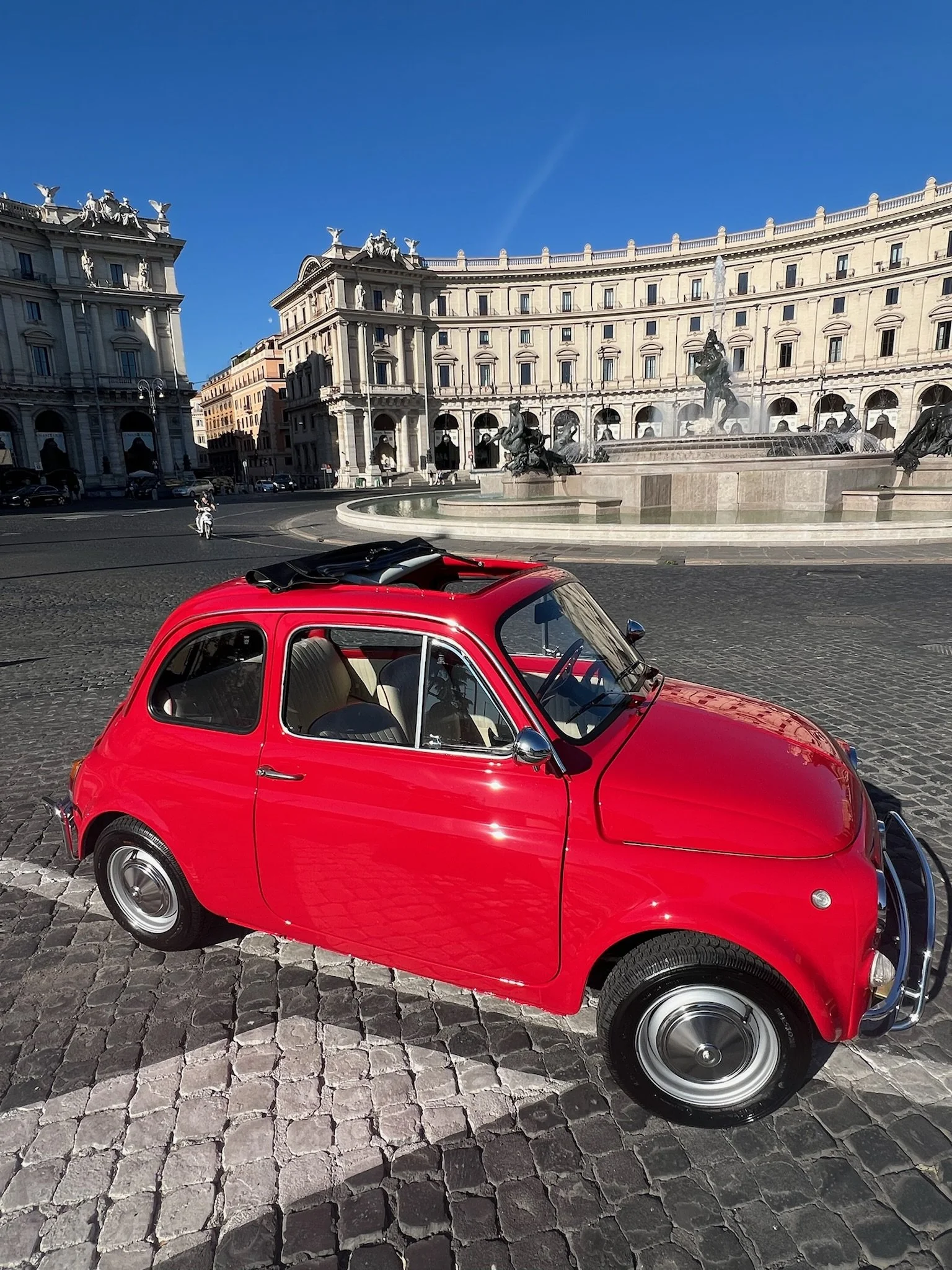 Una piccola auto rossa parcheggiata su una strada di ciottoli davanti a una fontana con sculture di cavalli e cavallieri, con edifici storici sullo sfondo e un cielo blu chiaro.