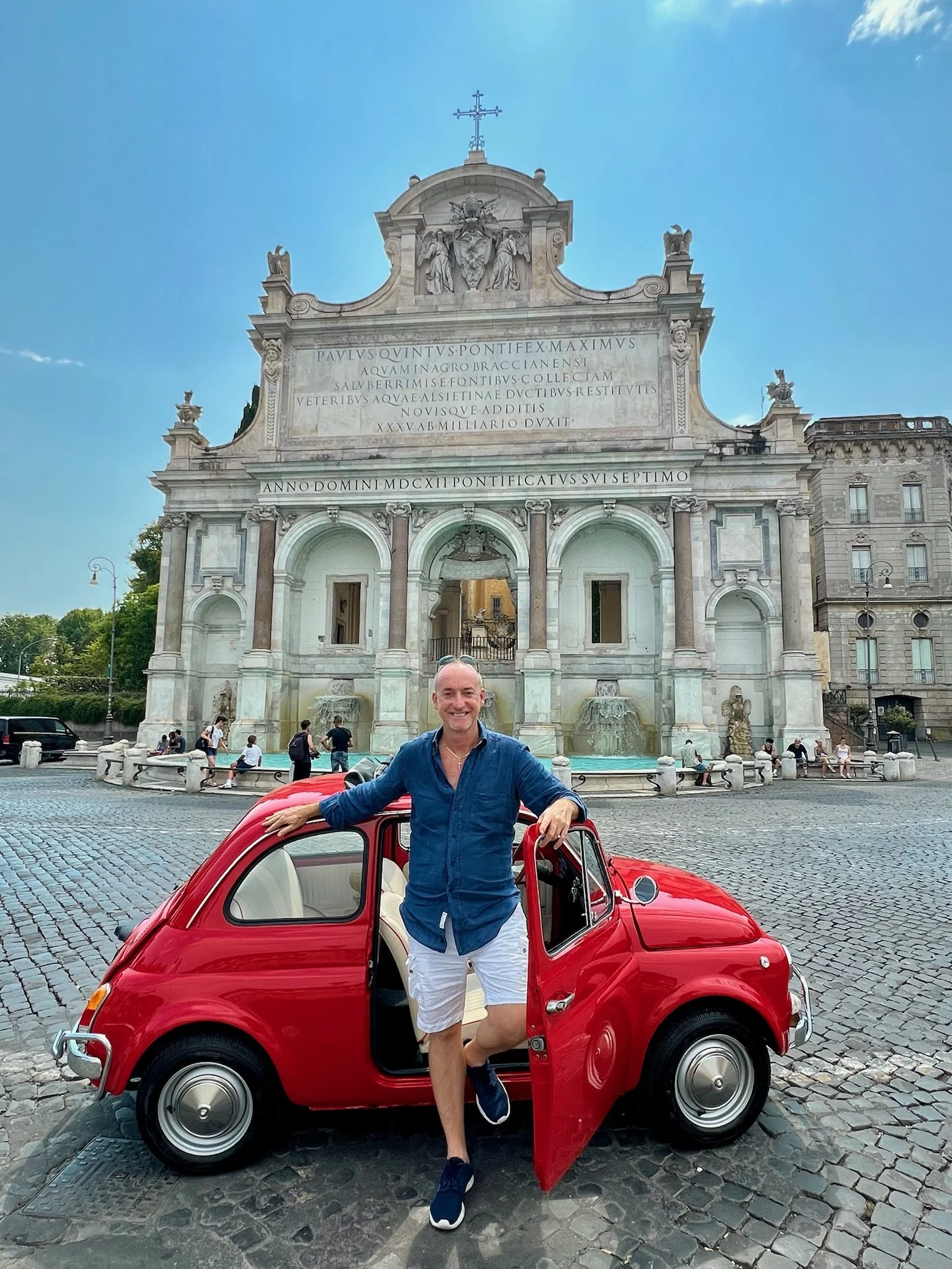 Uomo sorridente con camicia blu e pantaloncini bianchi si presenta accanto a una piccola auto rossa in Piazza della Fontana di Trevi, Roma, con la Fontana di Trevi sullo sfondo un monumento storico in stile barocco.