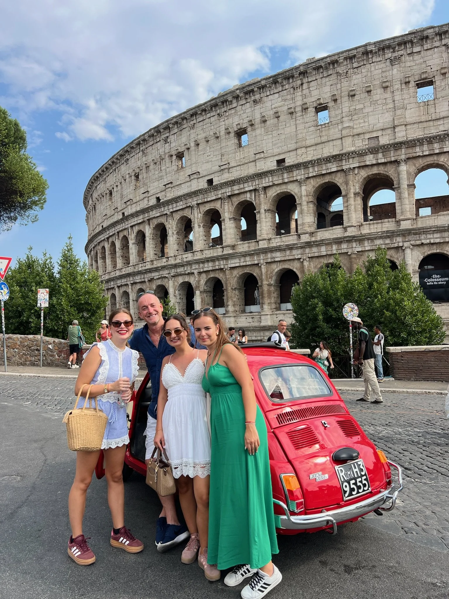 Gruppo di quattro persone sorridenti davanti a una piccola auto rossa, con il Colosseo sullo sfondo, in una giornata soleggiata a Roma.