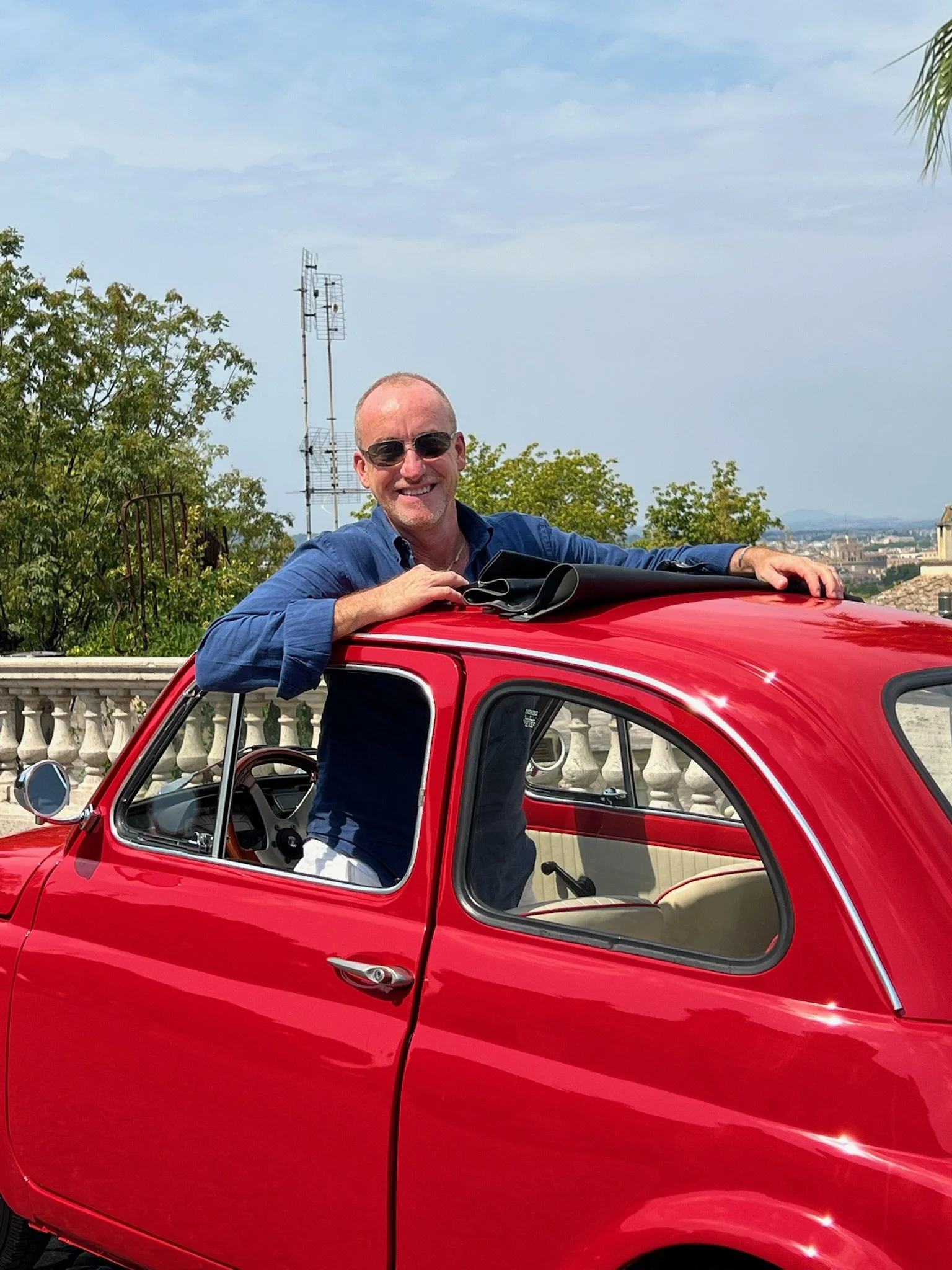 Uomo sorridente con occhiali da sole appoggiato sul tetto di una piccola auto rossa, con un paesaggio urbano sullo sfondo.