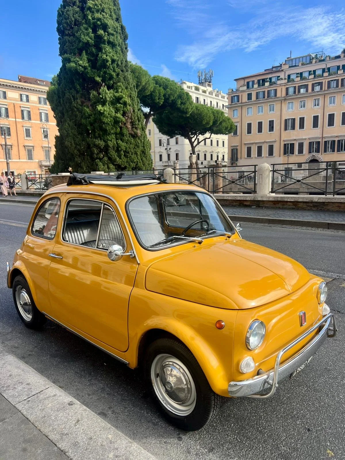 Una macchina gialla vintage parcheggiata lungo una strada con edifici e alberi sullo sfondo.