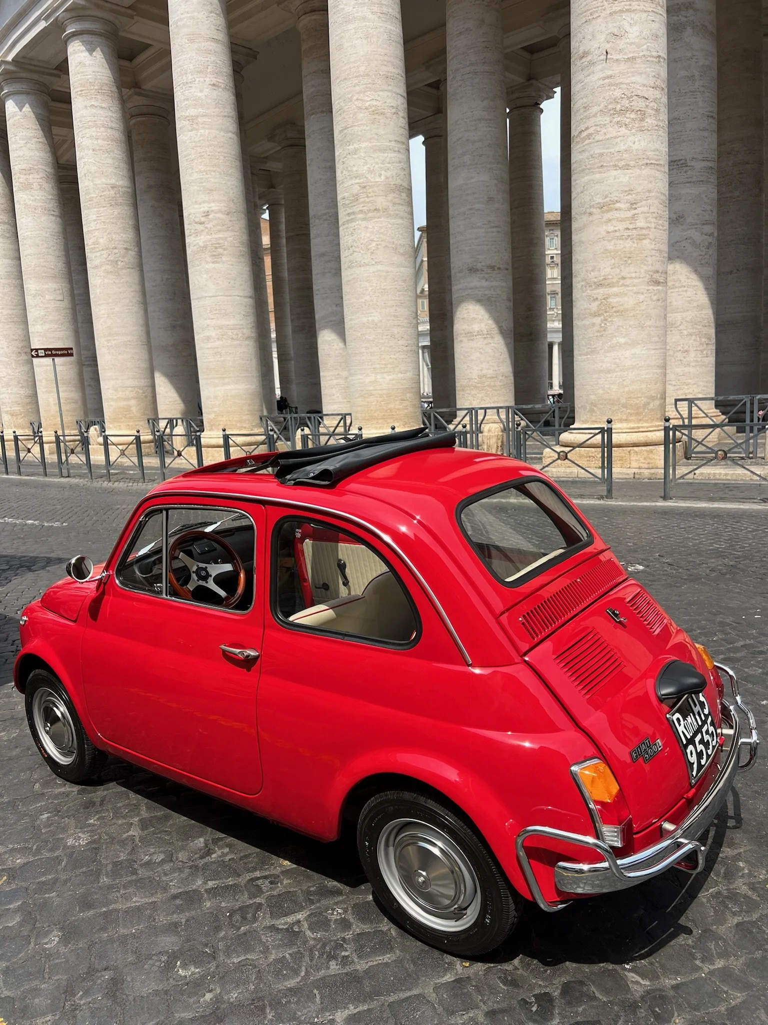 Fiat 500 rossa vintage parcheggiata su strada di pietra, con colonne di un edificio storico sullo sfondo.