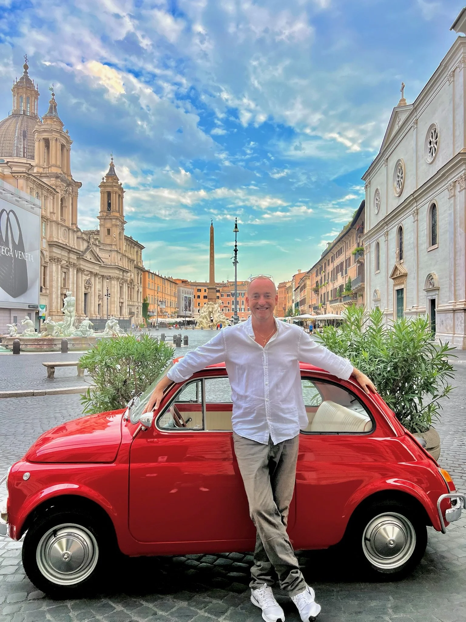 Un uomo sorridente si appoggia a una piccola auto rossa in una piazza di Roma con chiese antiche e un obelisco sullo sfondo, cielo azzurro con nuvole.