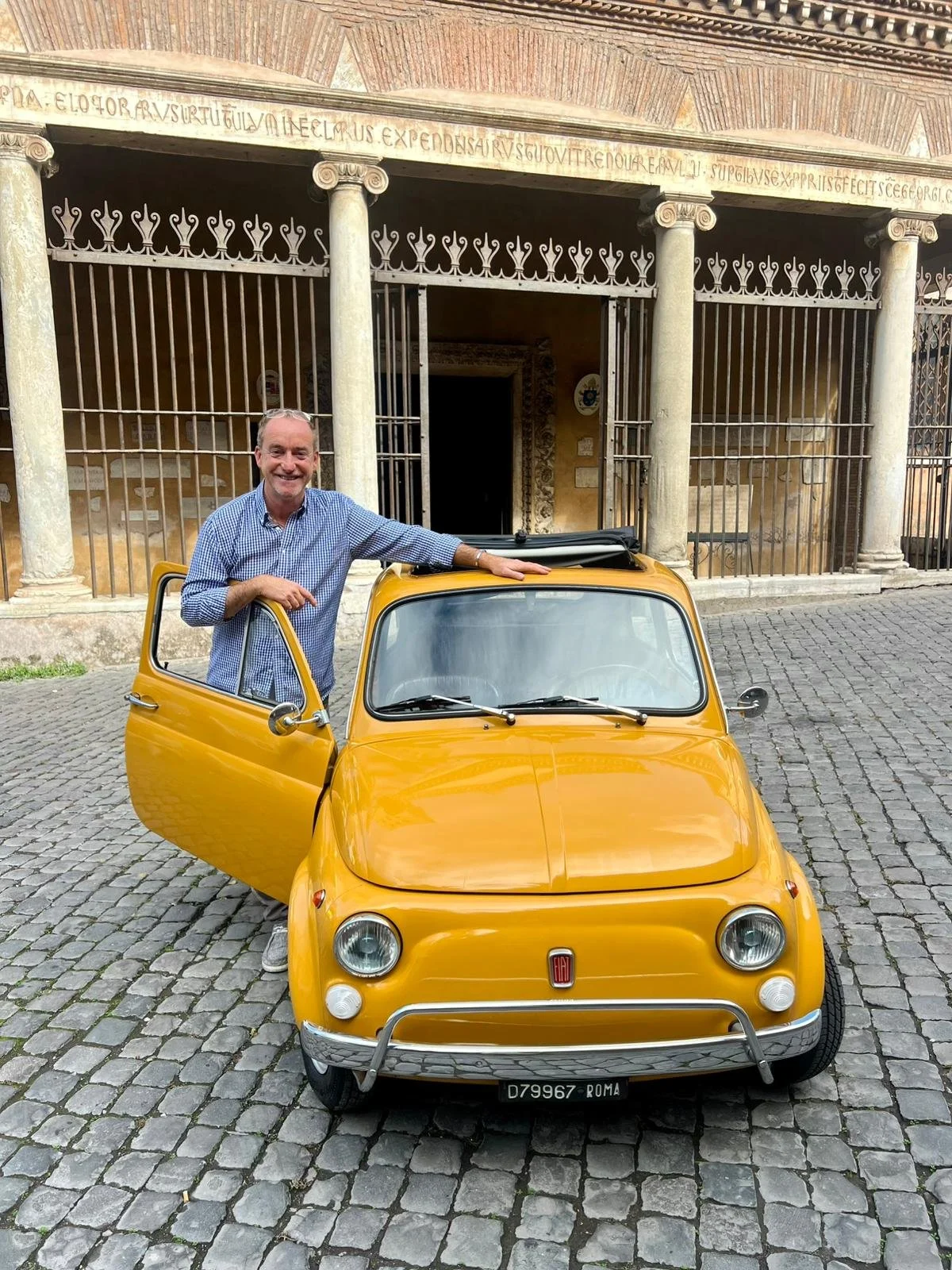 Uomo sorridente con camicia a quadri blu e pantaloni chiari che si appoggia sulla portiera aperta di un'auto gialla classica, Fiat 500, parcheggiata su una strada di ciottolato davanti a un edificio storico con colonne e iscrizioni latine sopra l'ing