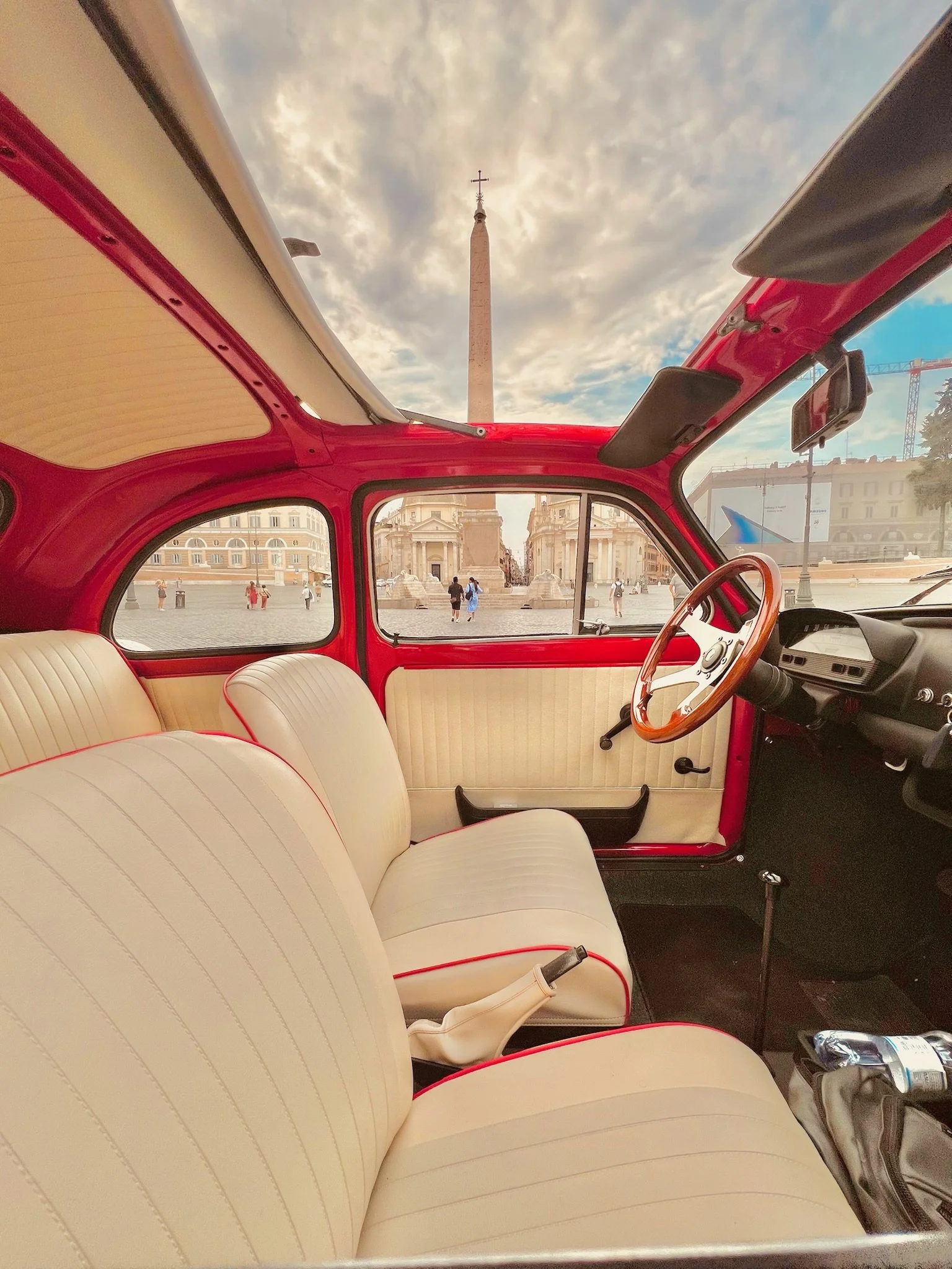 Interno di un'auto vintage con sedili beige e volante di legno, vista su piazza con obelisco nel centro di Roma, cielo nuvoloso.