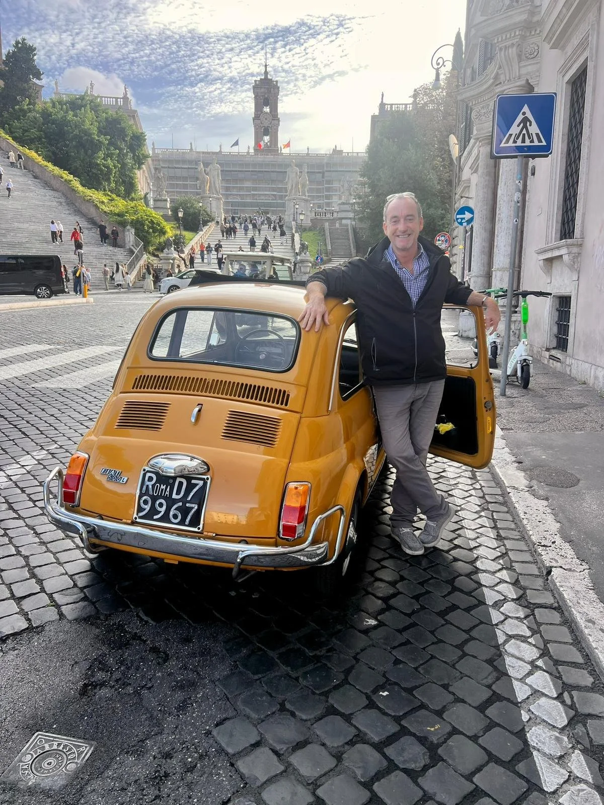 Uomo sorridente con giacca nera si appoggia a una piccola auto gialla in una zona cittadina con scalinate e monumenti sullo sfondo, cielo nuvoloso.