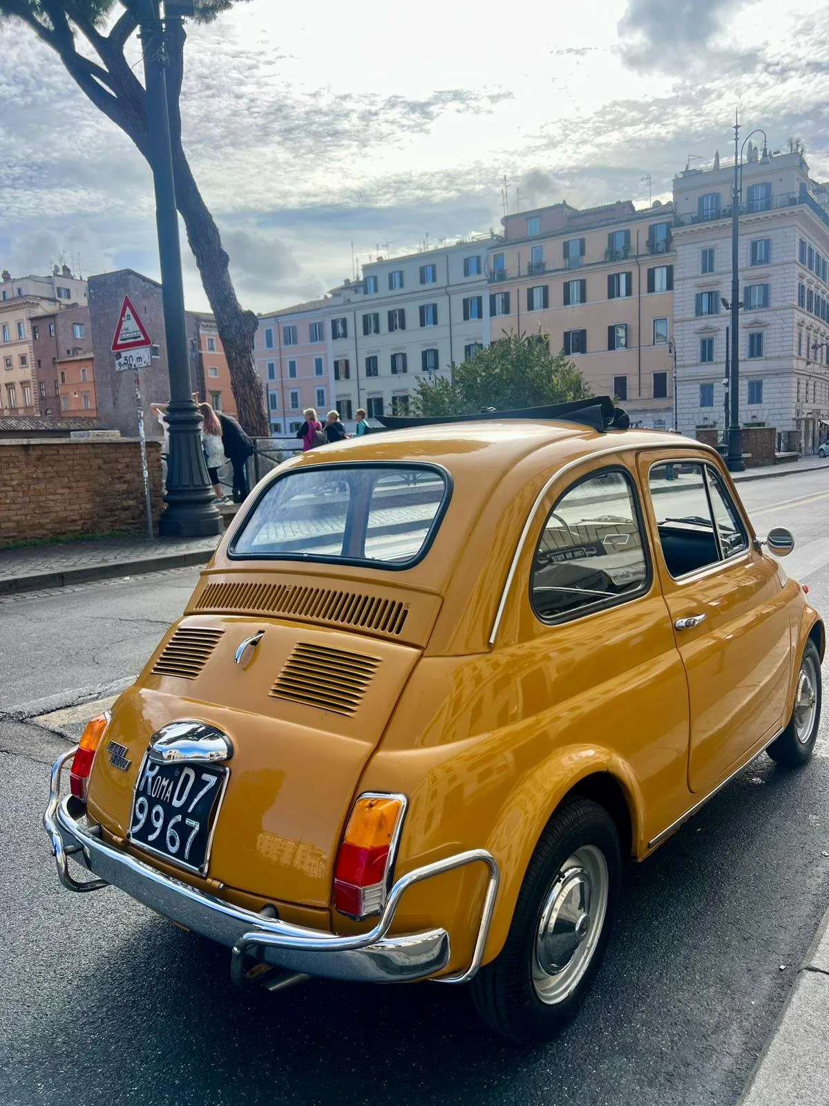 Auto vintage gialla parcheggiata su strada con edifici residenziali e un gruppo di persone in background, sotto un cielo nuvoloso.