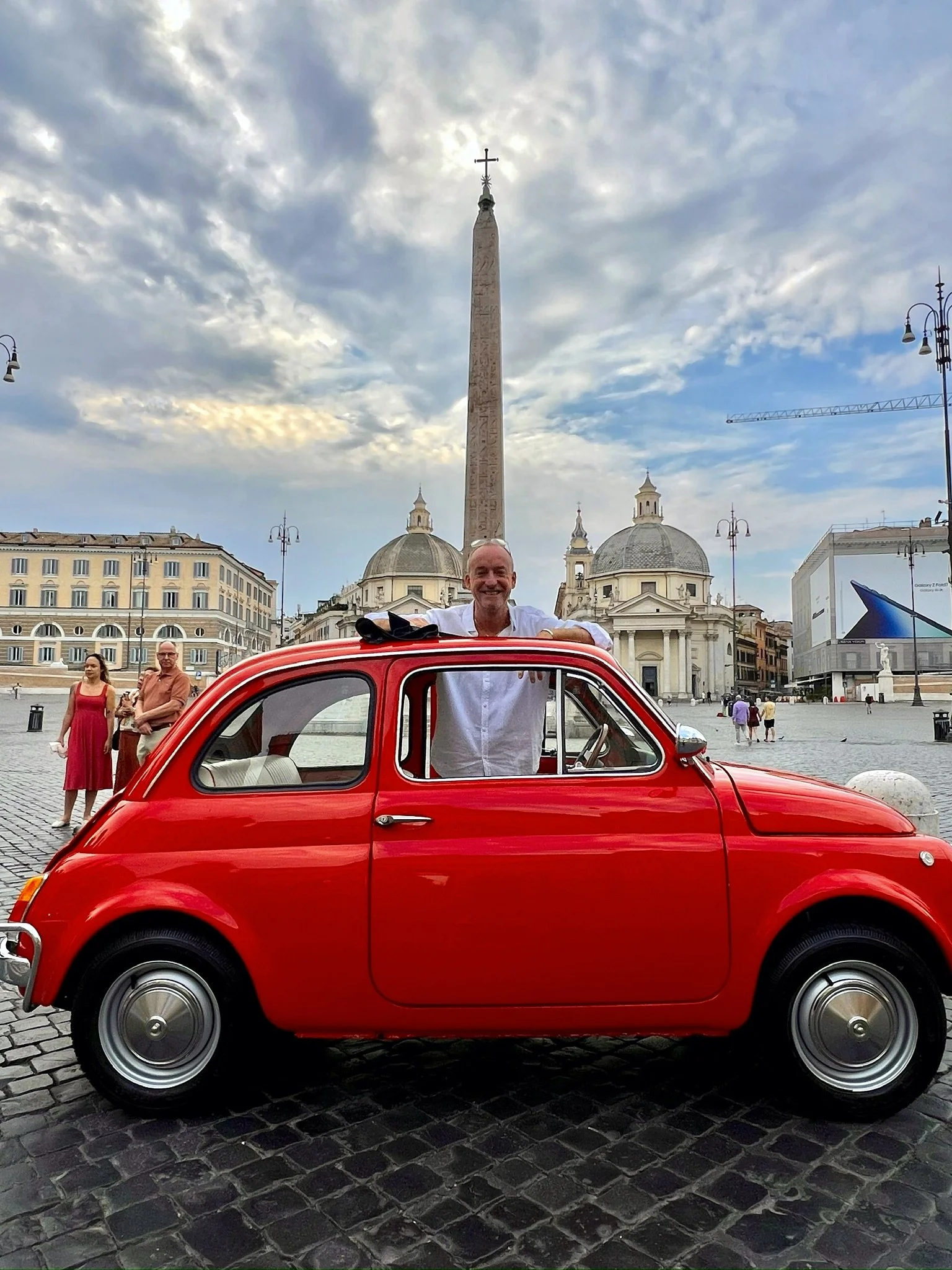 Uomo sorridente in una macchina rossa vintage Fiat 500, in Piazza del Popolo, Roma, con obelisco e chiese storiche sullo sfondo, cielo nuvoloso.