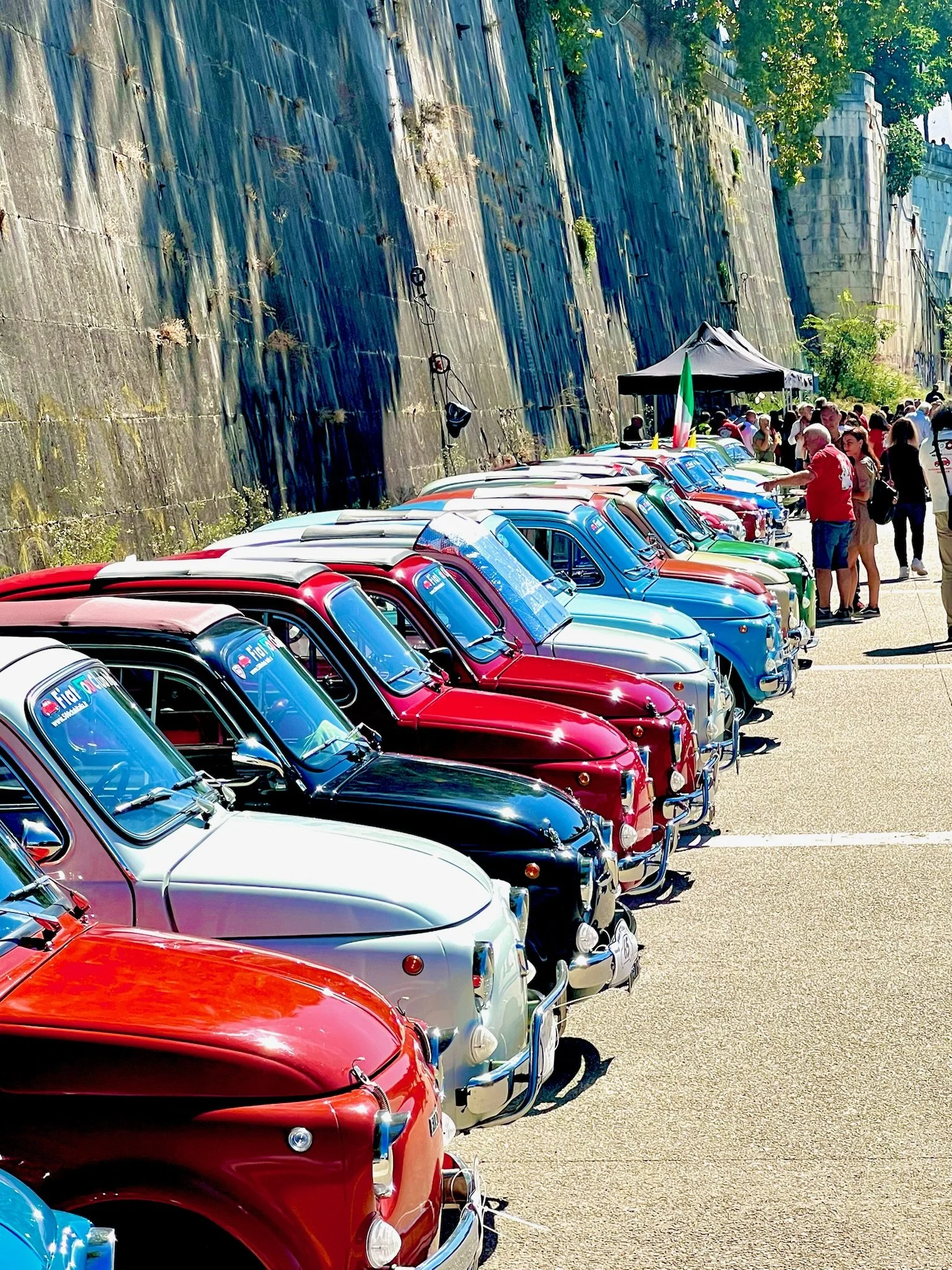 Raggruppamento di auto d'epoca di vari colori parcheggiate lungo un muro di pietra con persone che passeggiano e una tenda con bandiera italiana in background.
