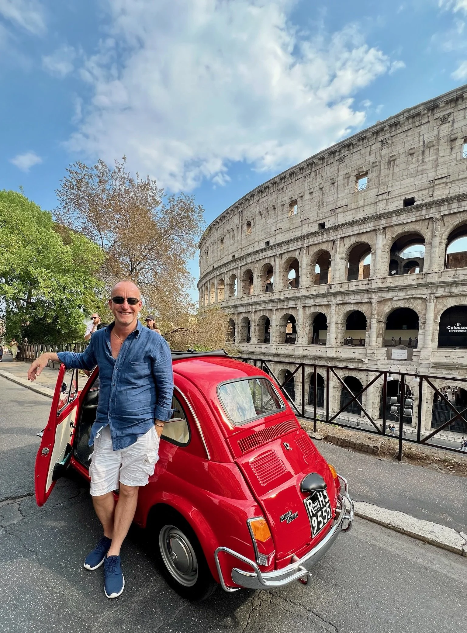 Un uomo felice con occhiali da sole, vestito con camicia blu e pantaloncini bianchi, si appoggia a una piccola auto rossa davanti al Colosseo a Roma, Italia, in una giornata di sole con alcune nuvole.