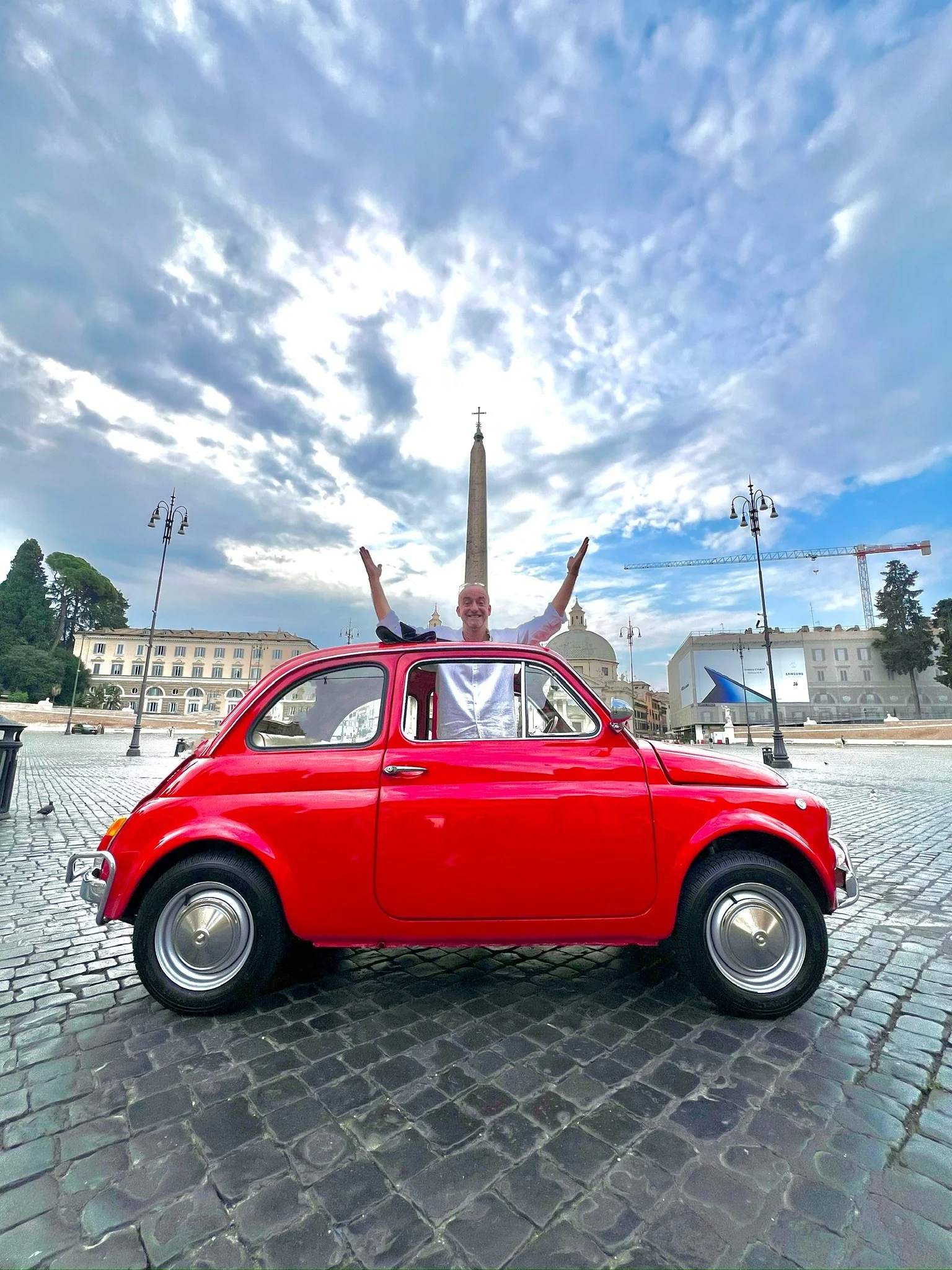 Uomo felice con braccia alzate sopra una macchina rossa San Giovanni in Laterano a Roma, con un obelisco al centro e un cielo nuvoloso.