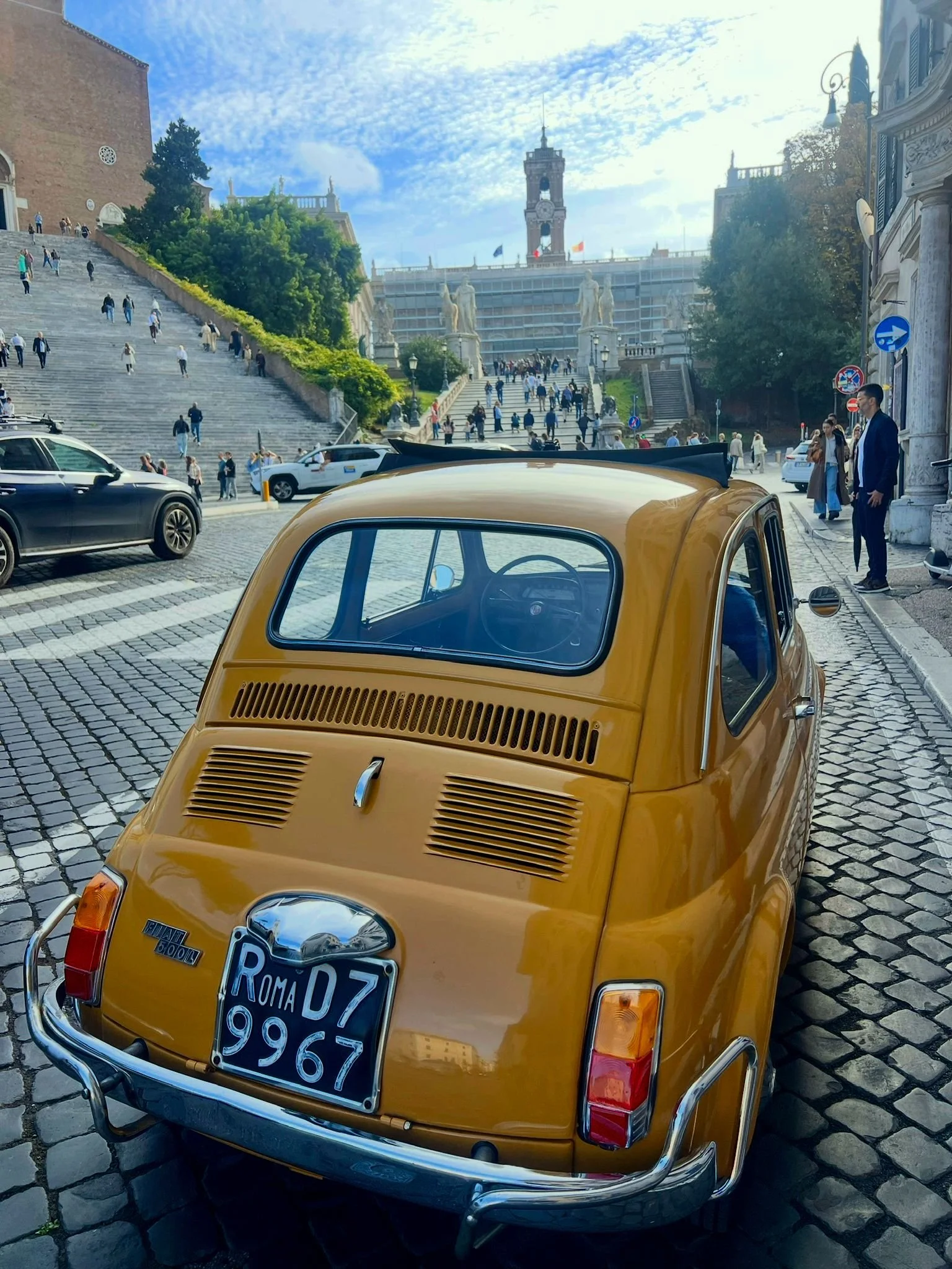 Una macchina gialla d'epoca parcheggiata su una strada di ciottoli, con le scale e una piazza affollata sullo sfondo, a Roma, Italia.