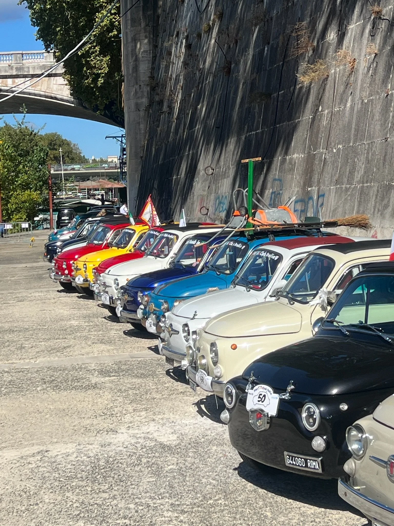 Una fila di auto d'epoca di vari colori parcheggiate lungo un muro in città, con un ponte visibile sullo sfondo e un cielo blu sereno.