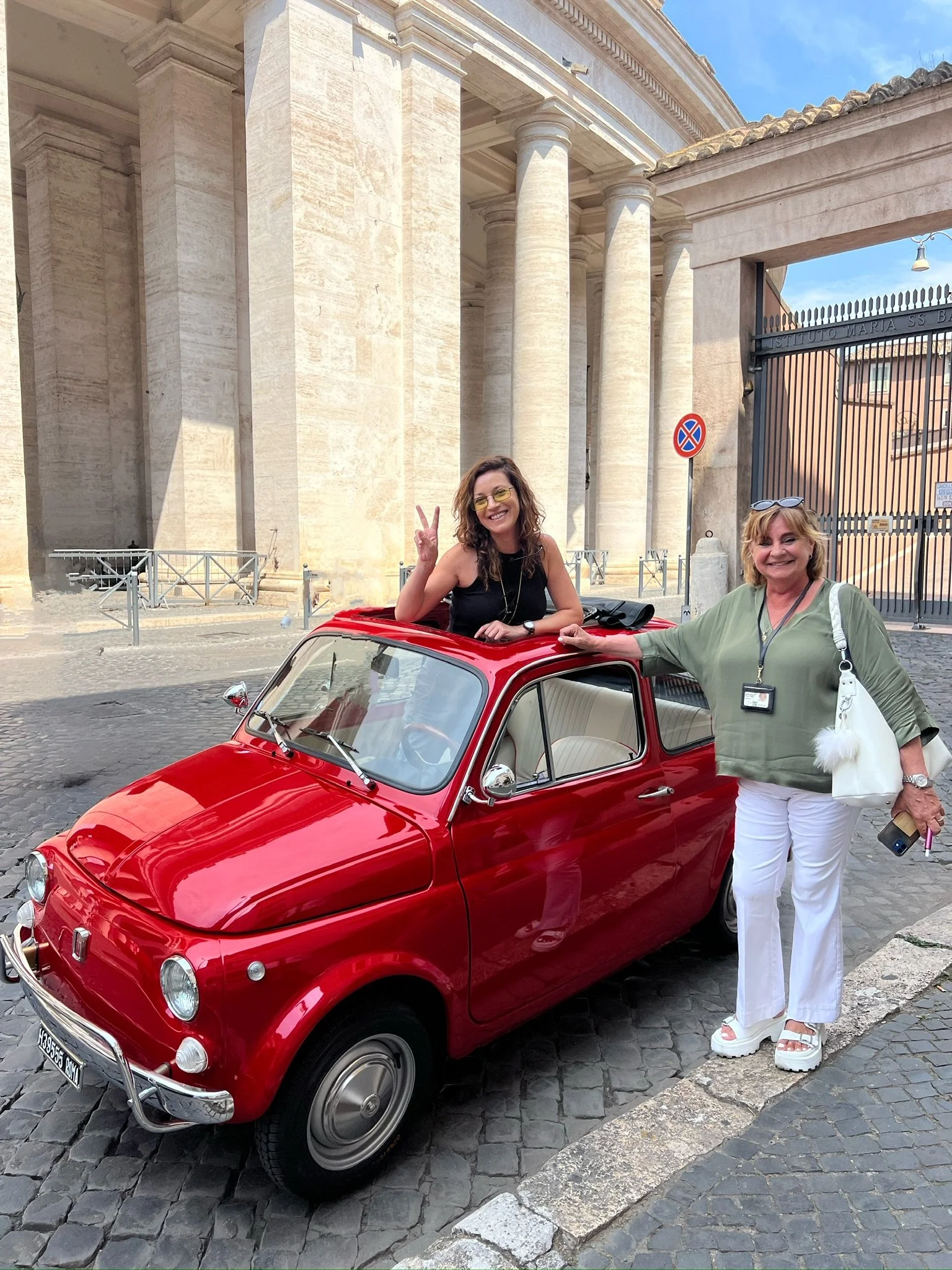Due donne sorridenti con una vettura rossa vintage davanti a un edificio storico con colonne, una donne sta in piedi, l'altra donna è seduta sul tetto dell'auto, entrambe fanno il gesto della pace.