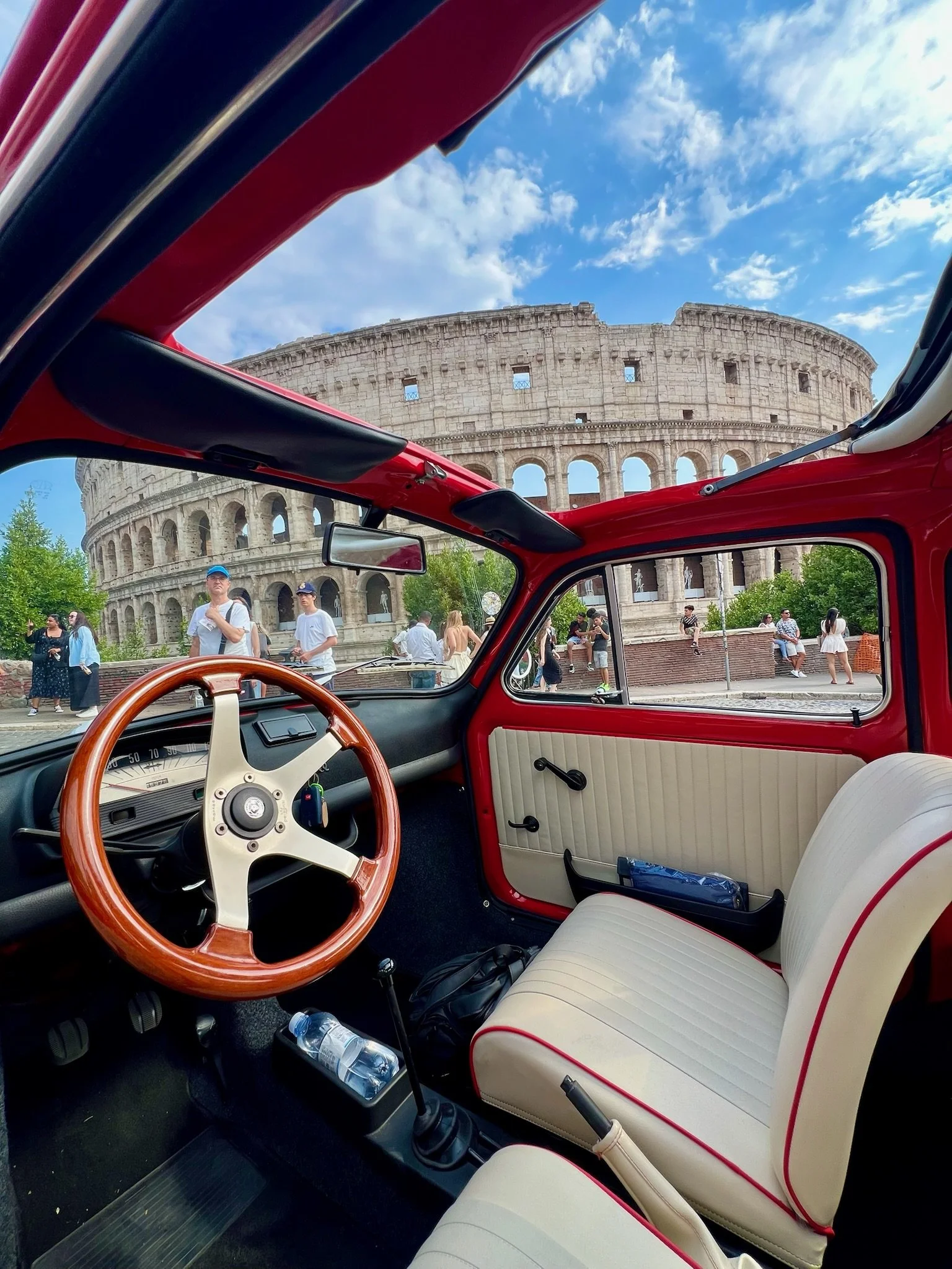 Interno di un'auto d'epoca rossa con volante in legno e sedili beige, con il Colosseo sullo sfondo in una giornata soleggiata con cielo azzurro e alcune nuvole.