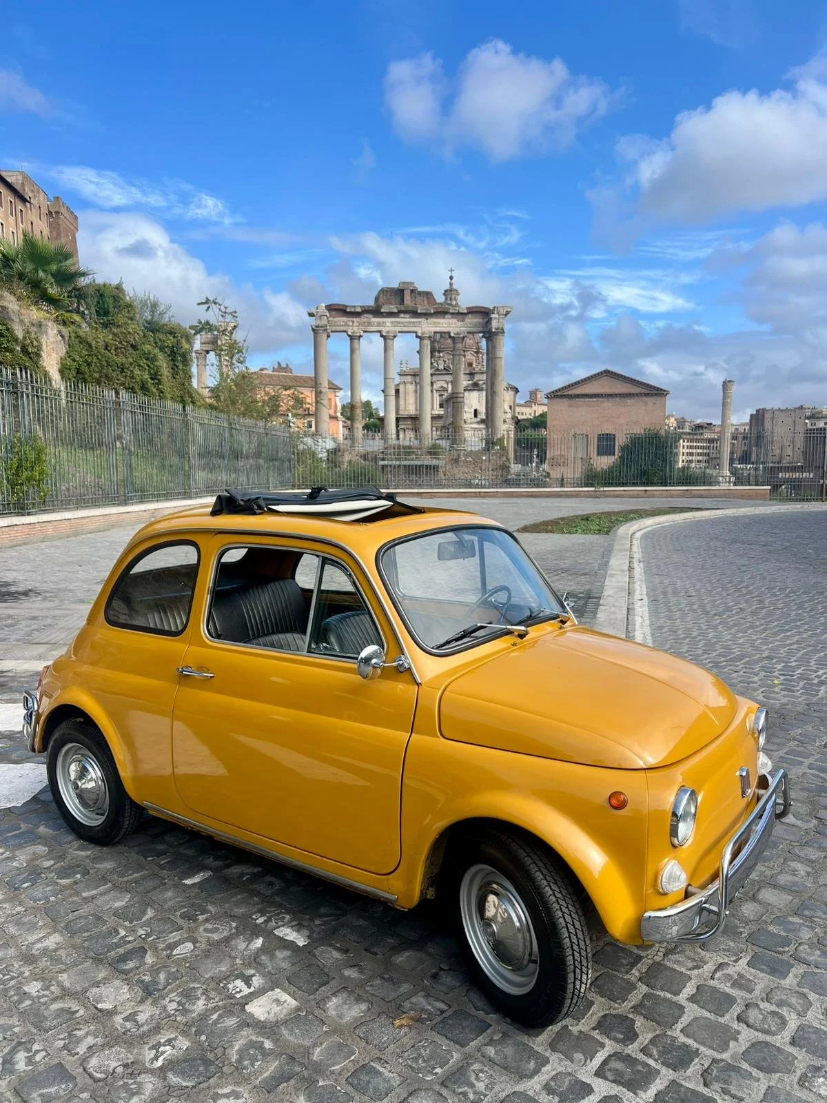 Macchina gialla vintage in strada con rovine romane e cielo nuvoloso sullo sfondo.