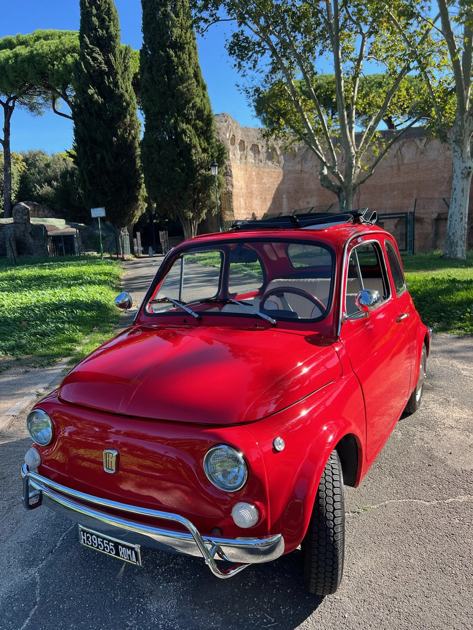 Auto d'epoca rossa parcheggiata su strada, con albero e mura antiche sullo sfondo.