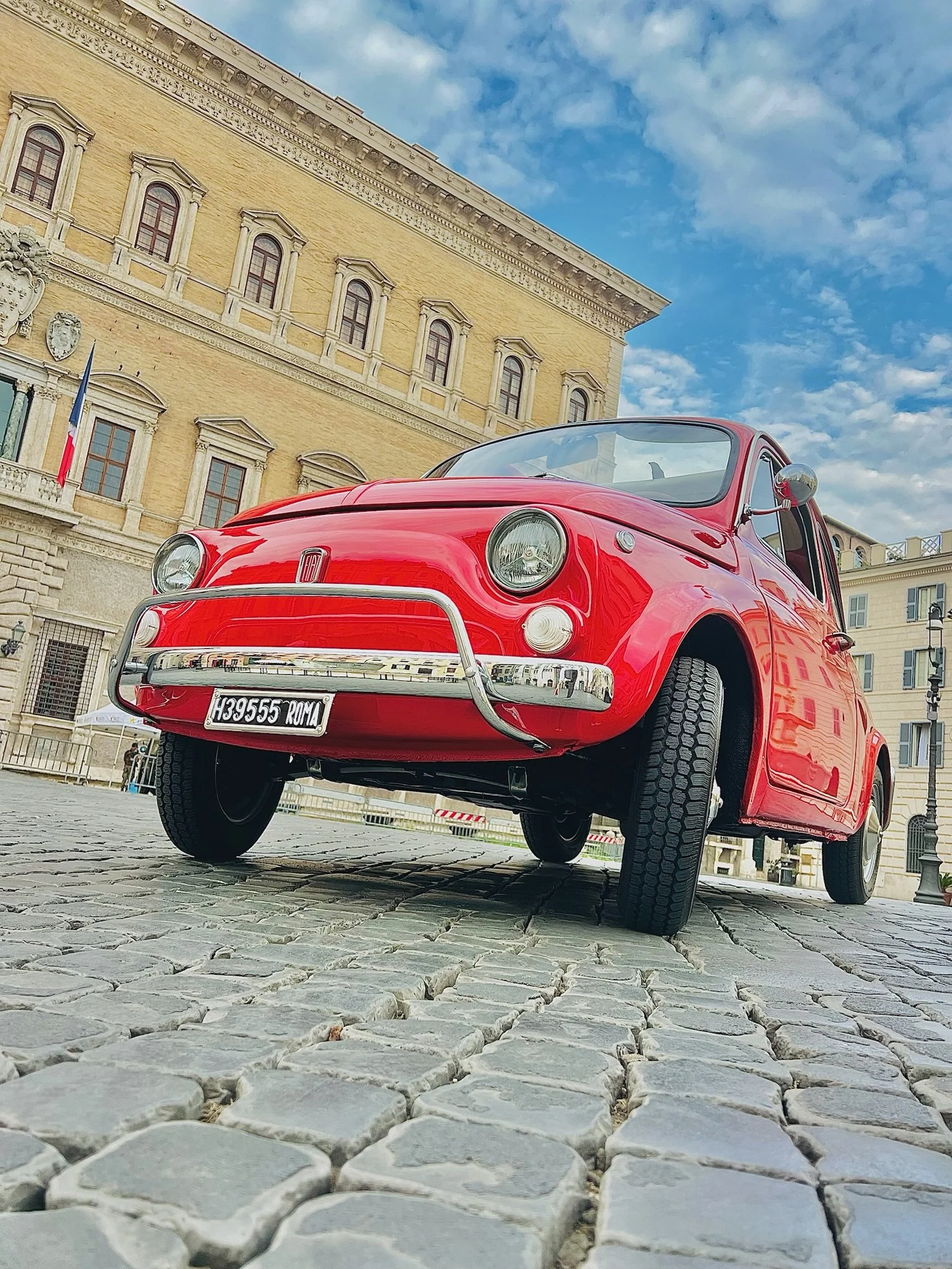Auto d'epoca rosso parcheggiato su una strada acciottolata con edifici storici e cielo azzurro sullo sfondo.