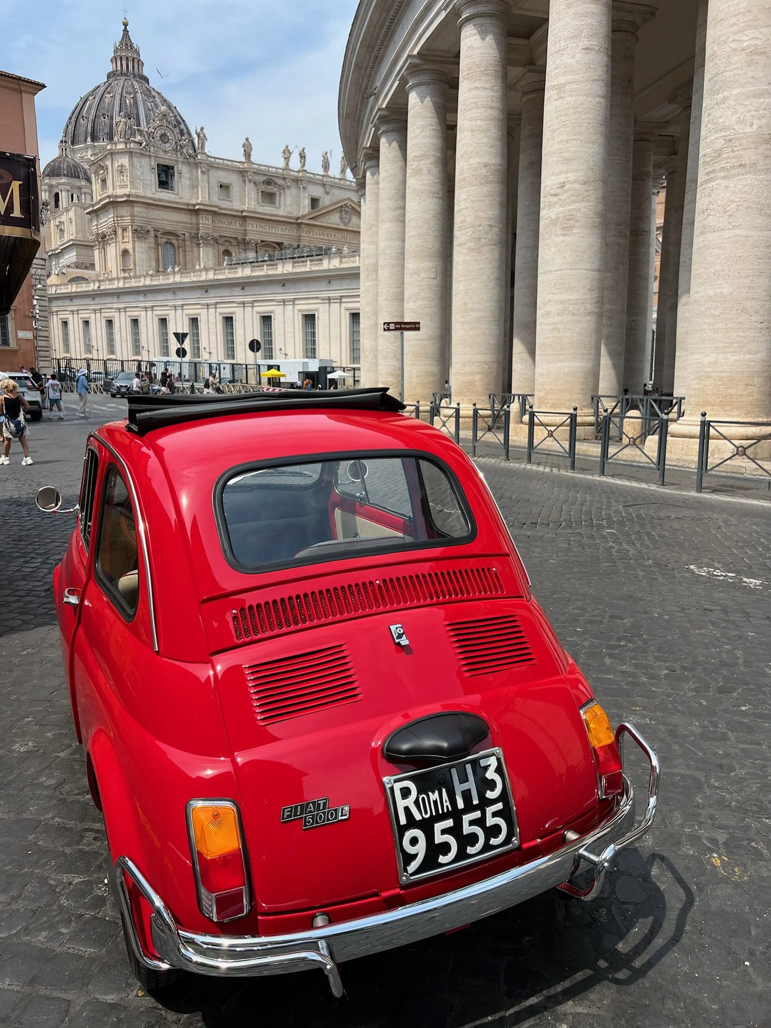Auto rossa Fiat 500L parcheggiata davanti a un colonnato storico con un edificio famoso di Roma sullo sfondo.