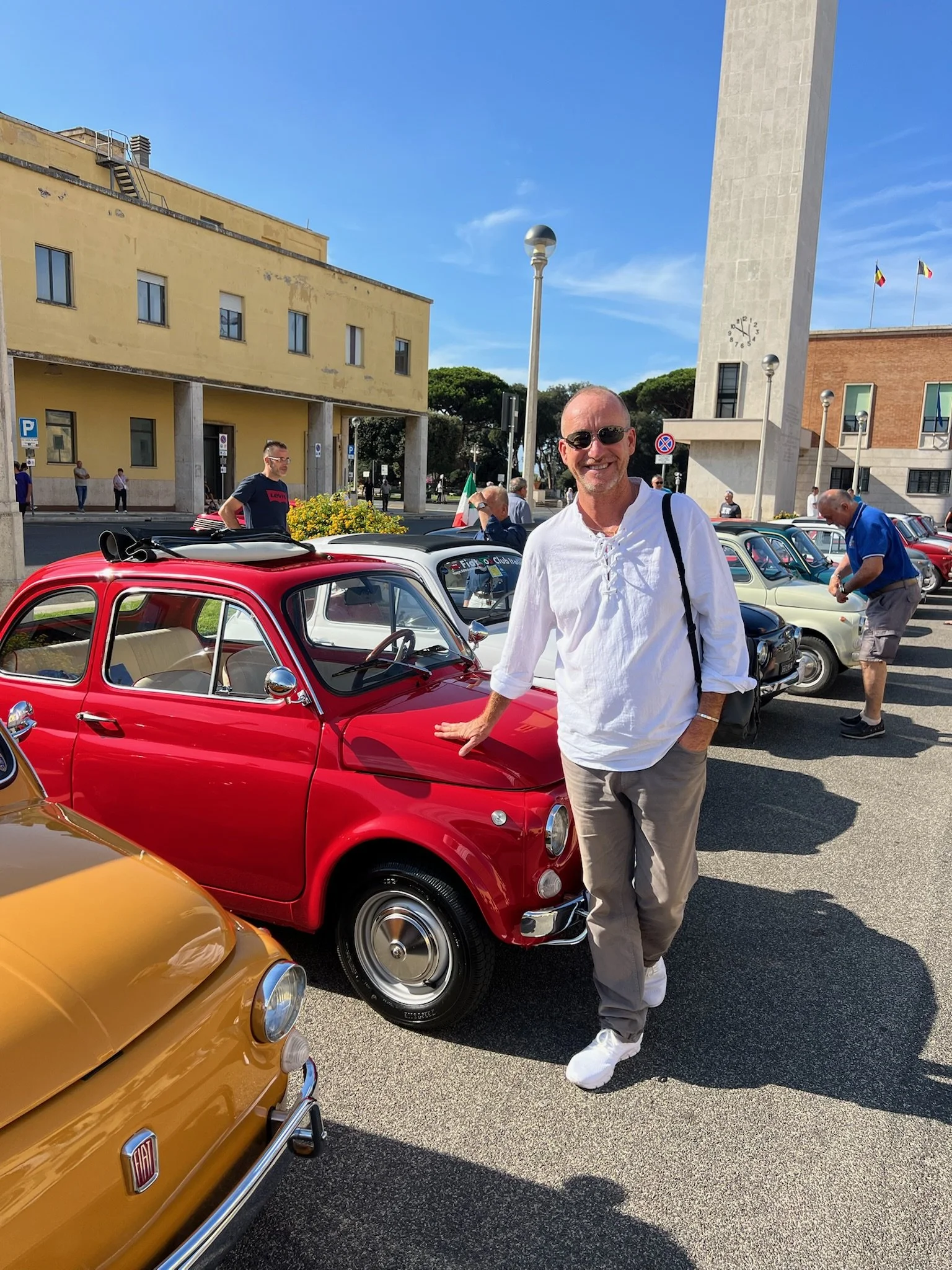 Uomo sorridente con occhiali da sole, vestito casual, che si appoggia a una piccola auto rossa in un evento di auto d'epoca, con altre auto d'epoca e persone intorno, sotto un cielo di giorno.