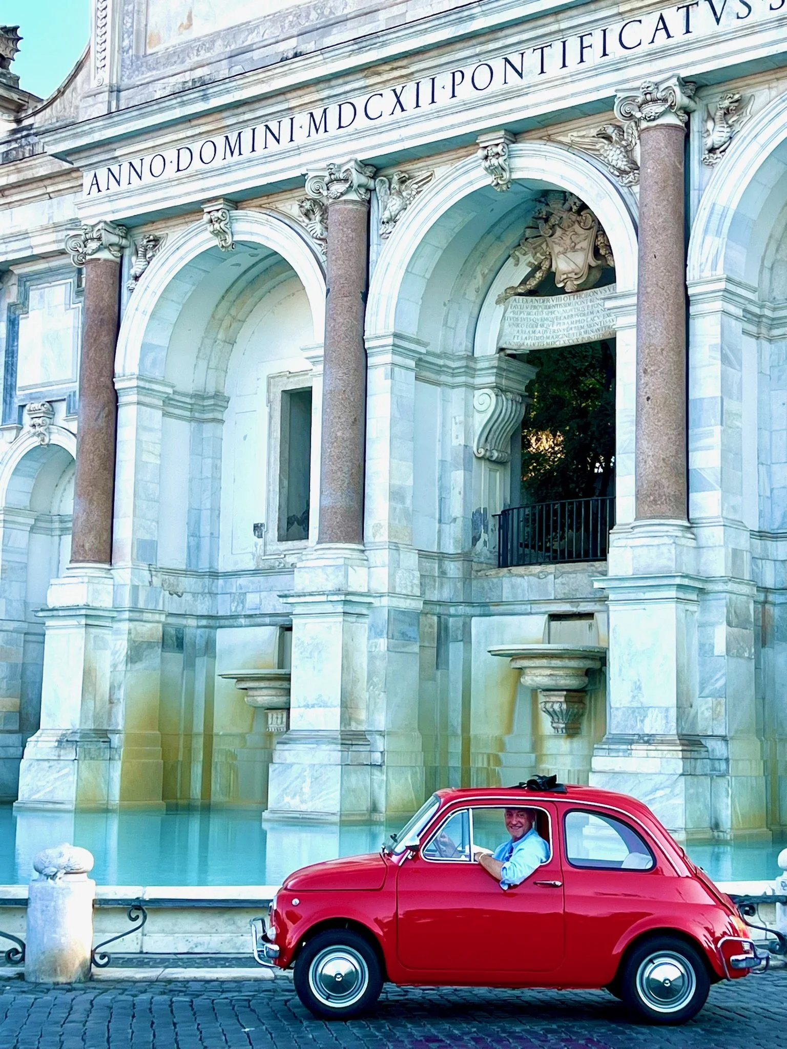 Una persona sorridente si trova alla guida di un'auto rossa di piccole dimensioni, parcheggiata davanti a un edificio storico in stile classico con archi e colonne.