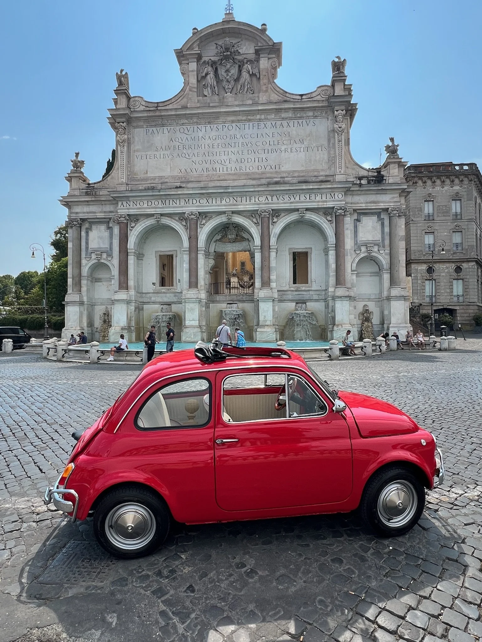 Una piccola automobile rossa parcheggiata su una strada di ciottoli davanti a una fontana e a un edificio storico in stile barocco con incisioni e sculture sulla facciata.