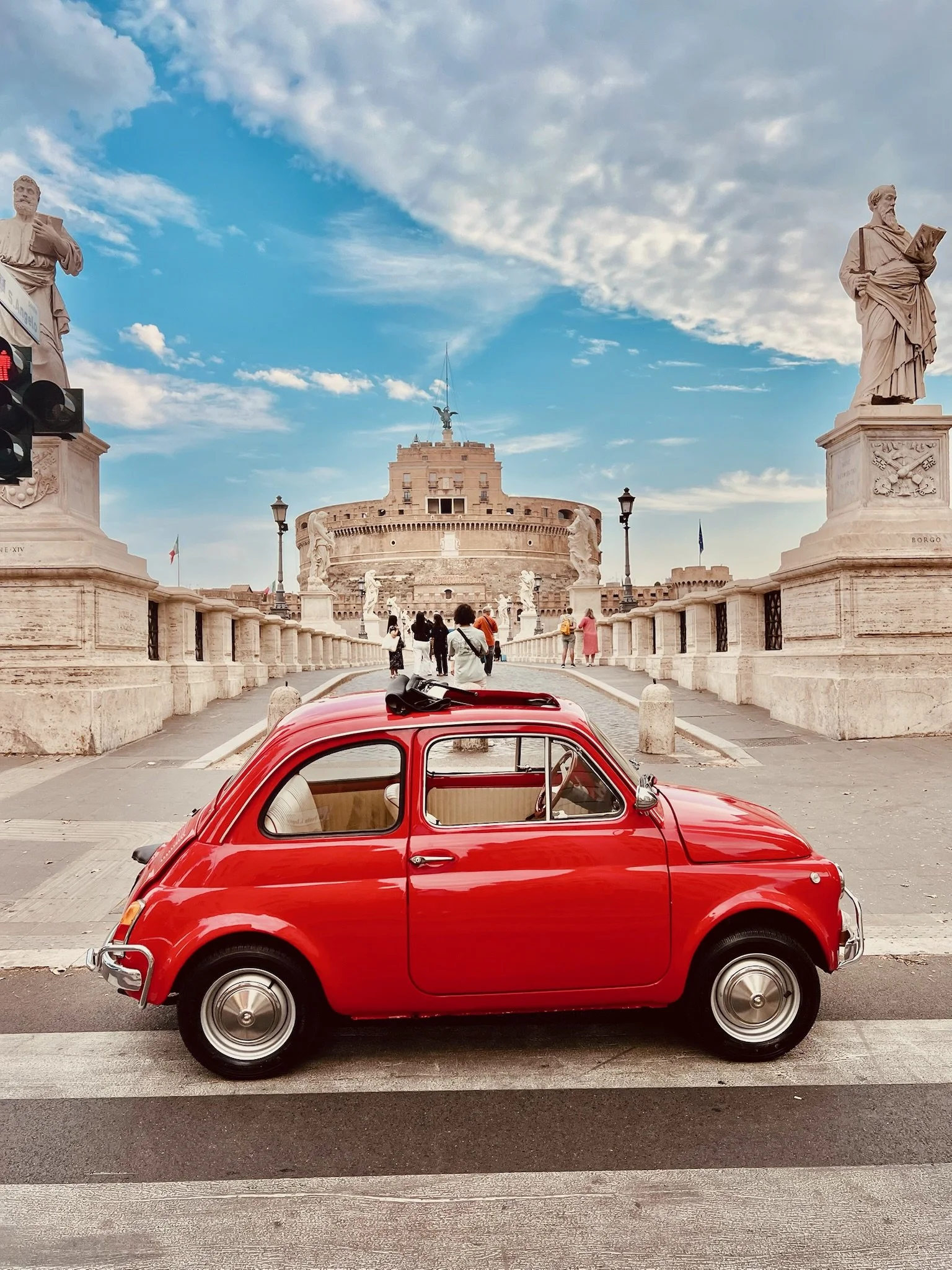 Auto rosso classico di fronte a Castel Sant'Angelo a Roma, con statue e persone sul ponte.