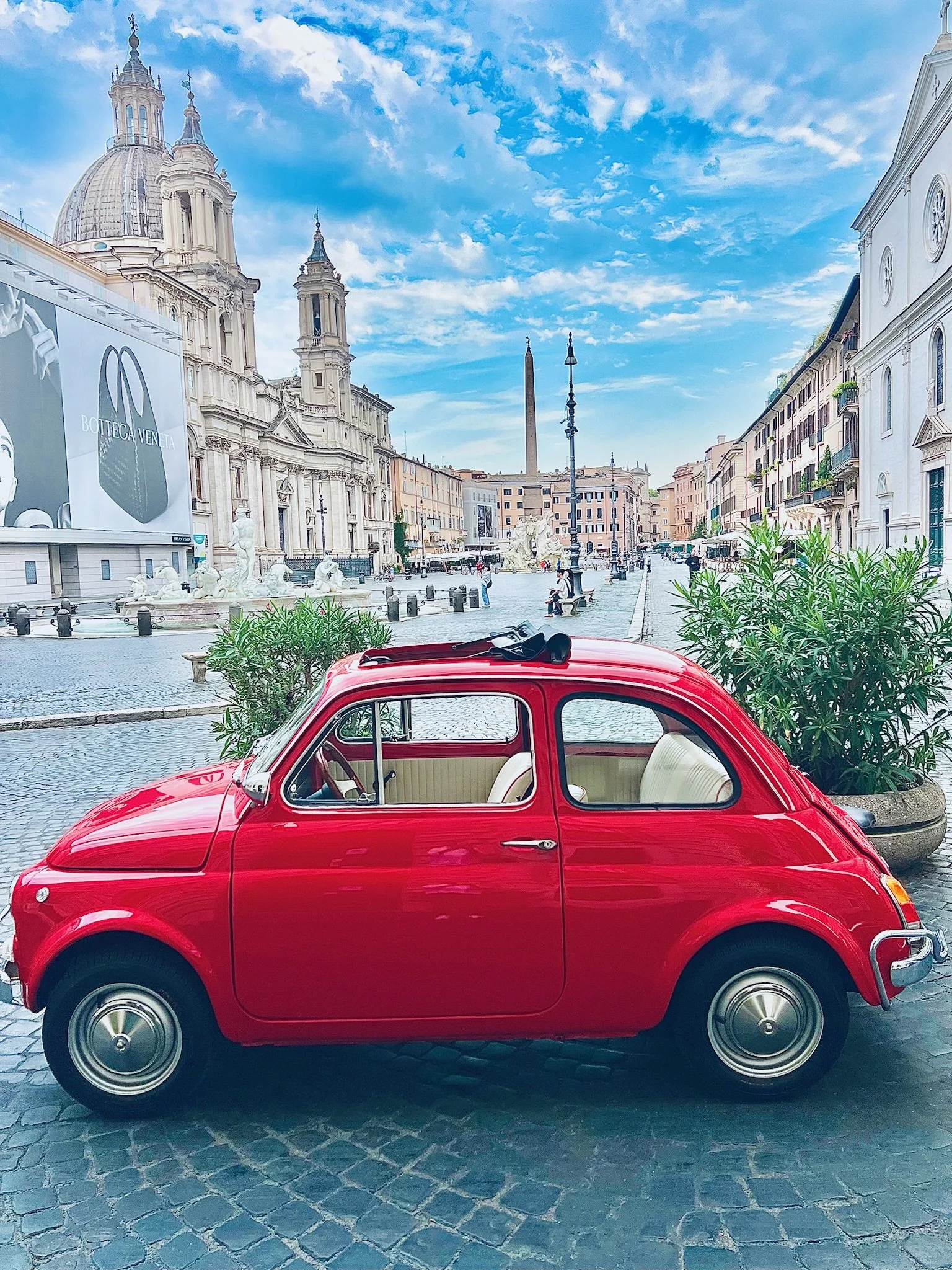 Una macchina rossa in stile vintage parcheggiata su una piazza con edifici storici, una chiesa con cupola e un obelisco, sotto un cielo azzurro con nuvole sparse.