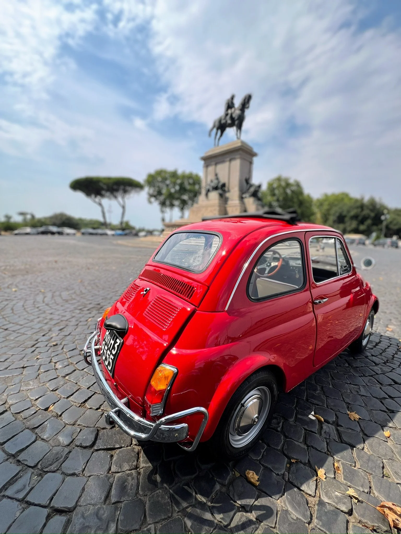Auto rosso in miniatura parcheggiato su una strada in pietra con statua di un cavaliere su cavallo sfondo e cielo con alcune nuvole
