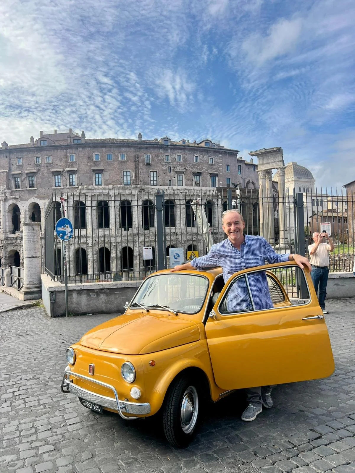 Uomo sorridente con camicia a quadri azzurro stando accanto a un'auto gialla vintage con la porta aperta, sullo sfondo si vede un antico sito archeologico e un uomo che scatta foto.