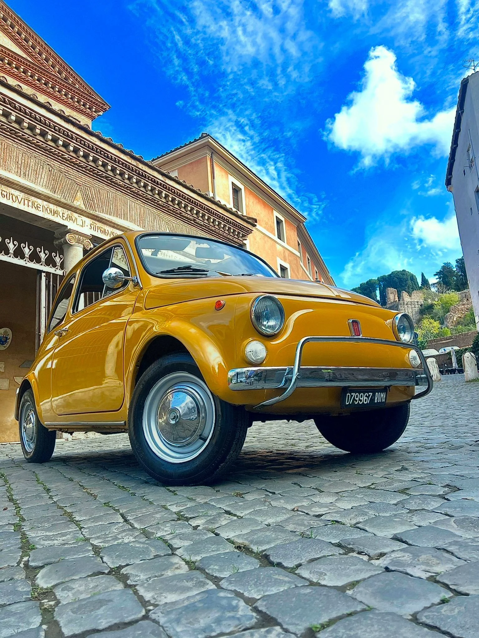 Auto d'epoca gialla parcheggiato su una strada in pietra, con edifici storici sullo sfondo e un cielo azzurro con nuvole sparse.
