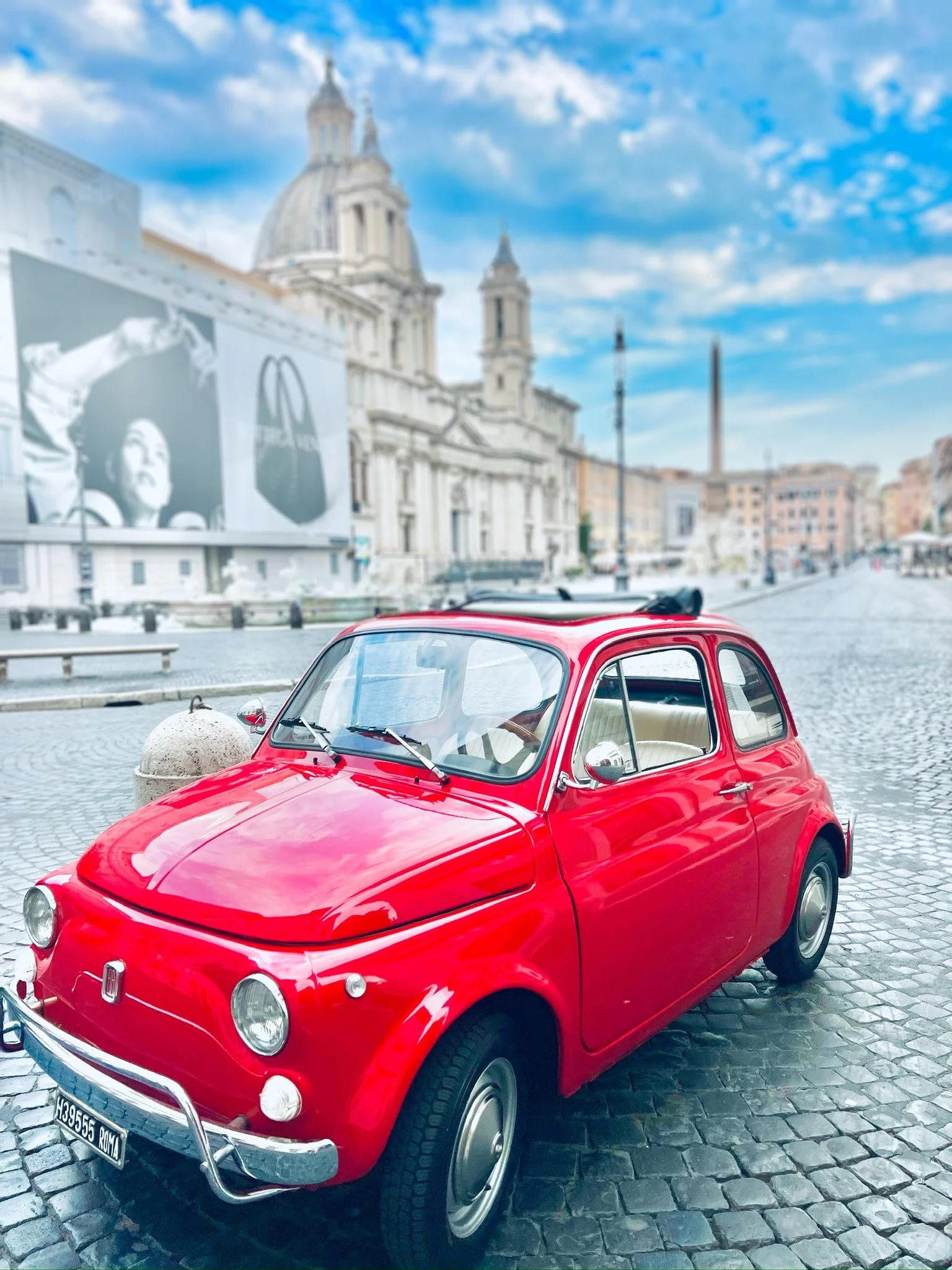 Una piccola auto rossa davanti a un edificio storico con una grande chiesa dal soffitto arcuato, un cielo blu con nuvole e una pubblicità di un negozio di moda in fondo.