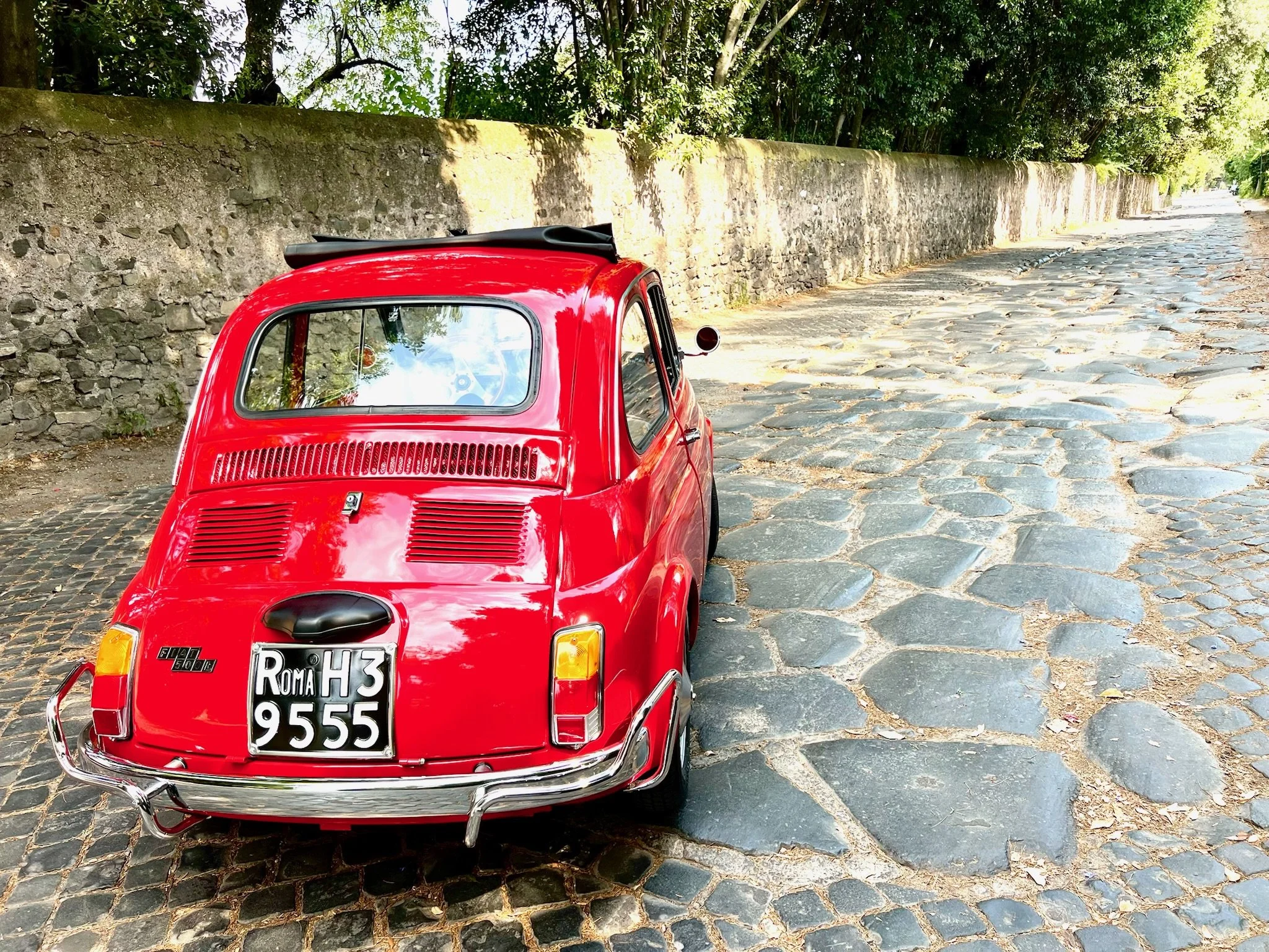 Auto rossa vintage parcheggiata su strada in pietra, con muro e alberi sullo sfondo.