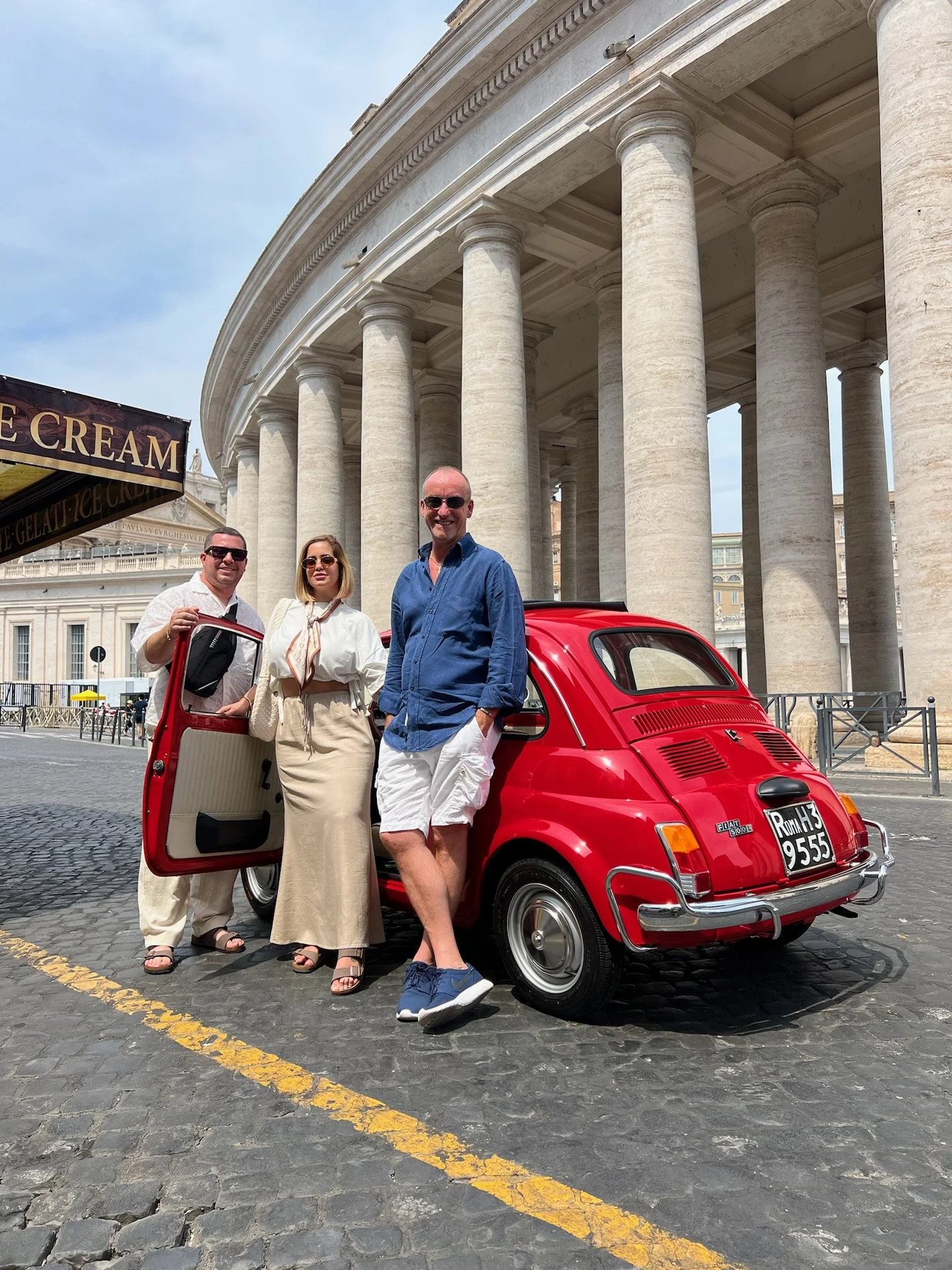 Tre persone sorridenti posano davanti a una piccola auto rossa in una piazza con colonne in stile classico. La scena è all'aperto durante il giorno.