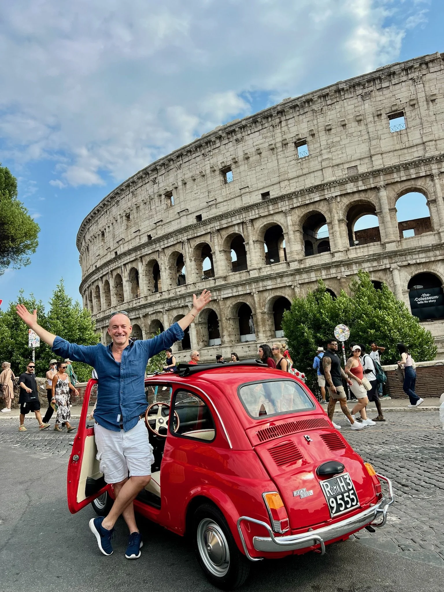 Un uomo sorridente con braccia alzate davanti a un'auto rossa in stile vintage, con il Colosseo sullo sfondo a Roma, Italia.
