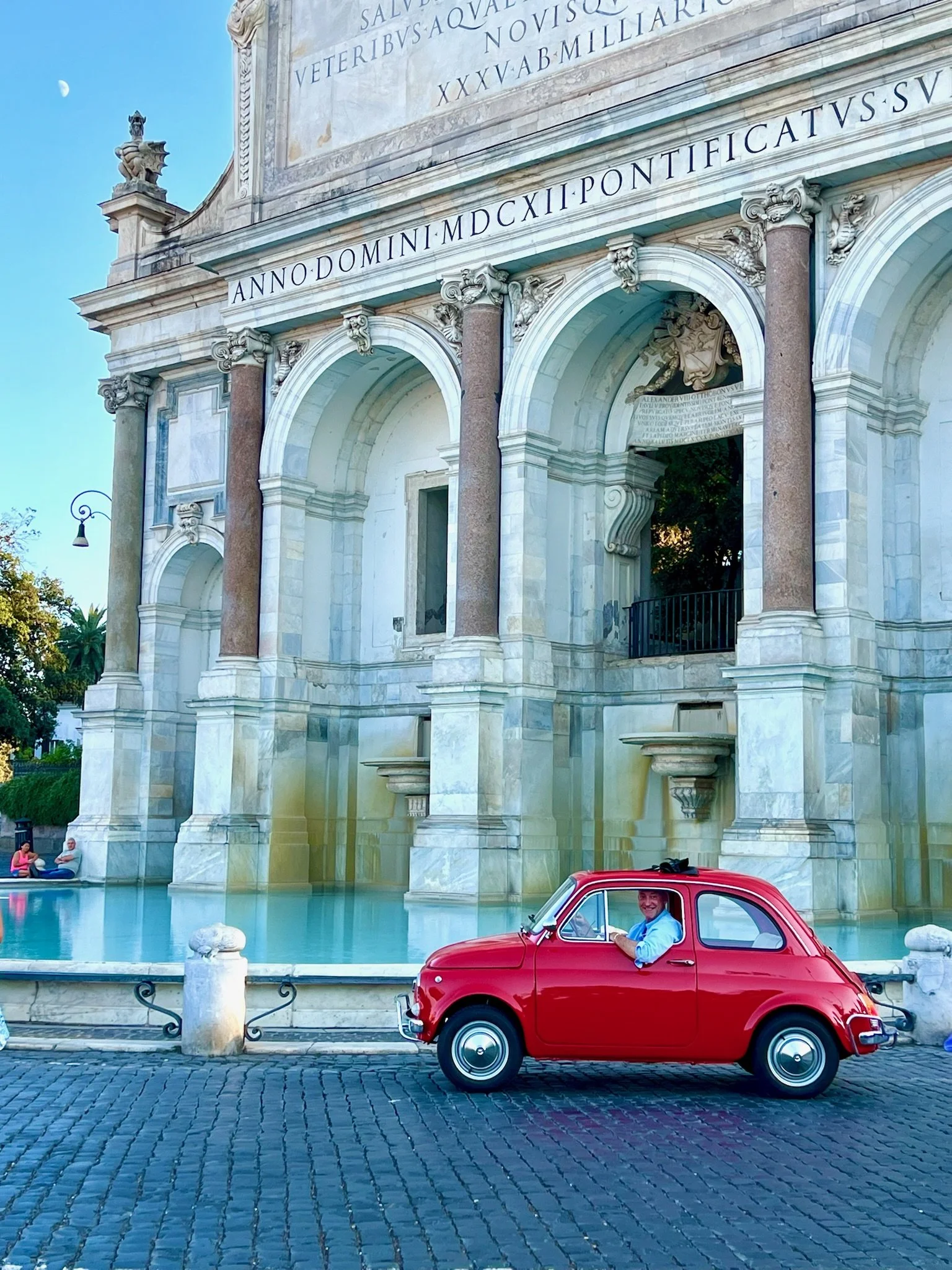 Un uomo sorridente seduto in un'auto rossa vintage davanti a un imponente edificio storico in stile classico, con colonne e iscrizioni in latino, un fontane e un cielo chiaro.