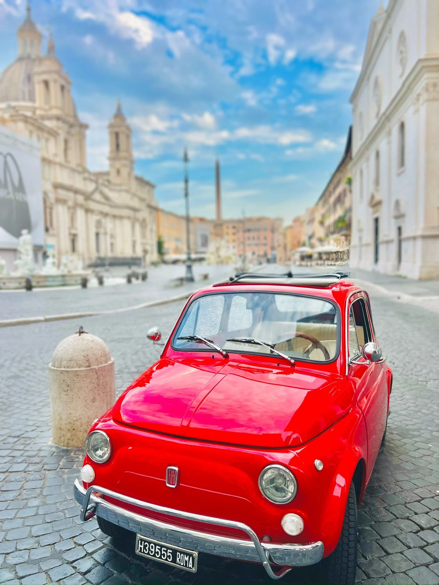 Auto d'epoca rossa parcheggiata su una strada di ciottoli con edifici storici e cielo azzurro con nuvole sparse sullo sfondo.