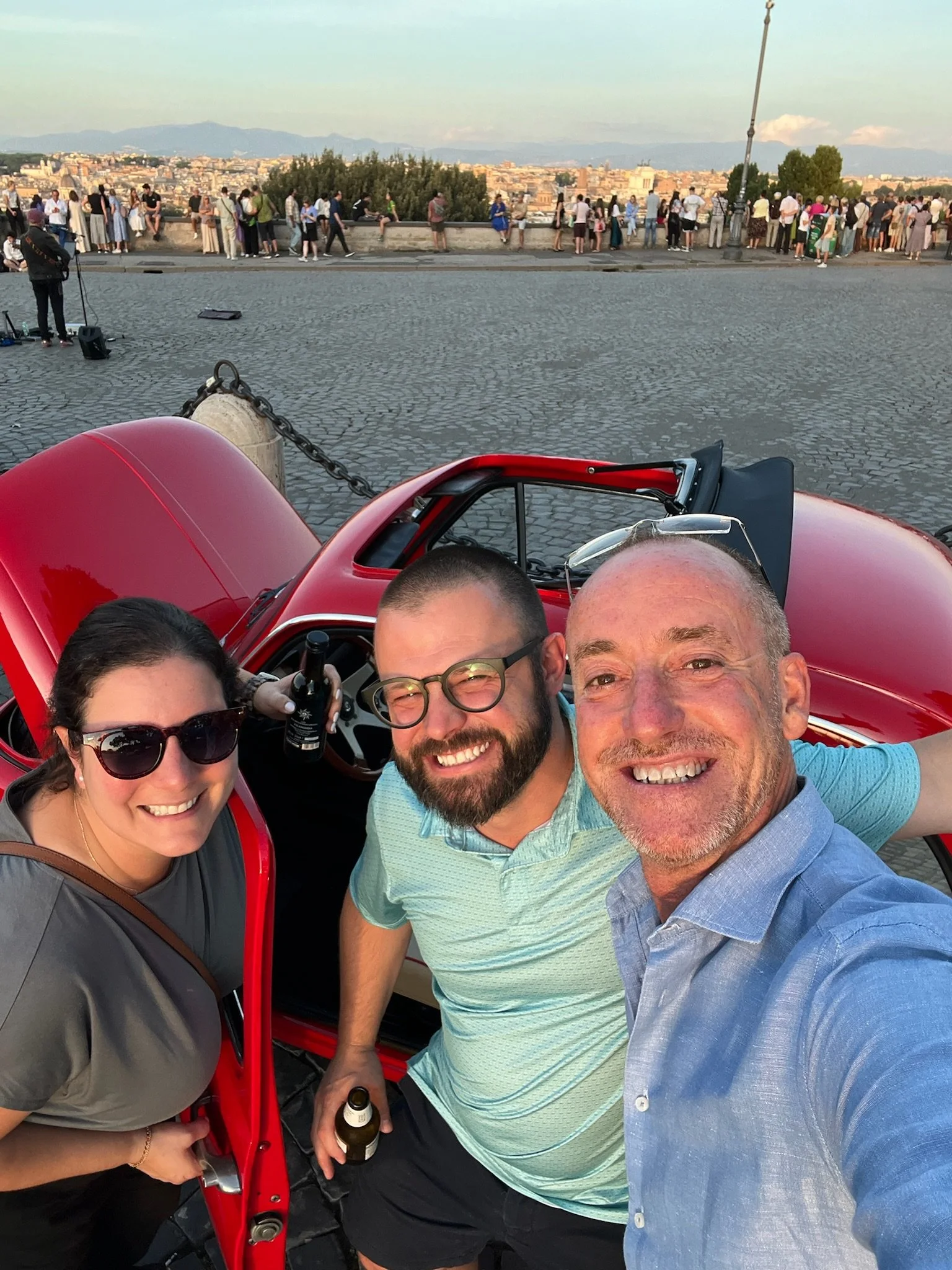 Tre persone felici che si scattano un selfie davanti a una piccola macchina rossa con una vista panoramica di una città e molte persone sullo sfondo.