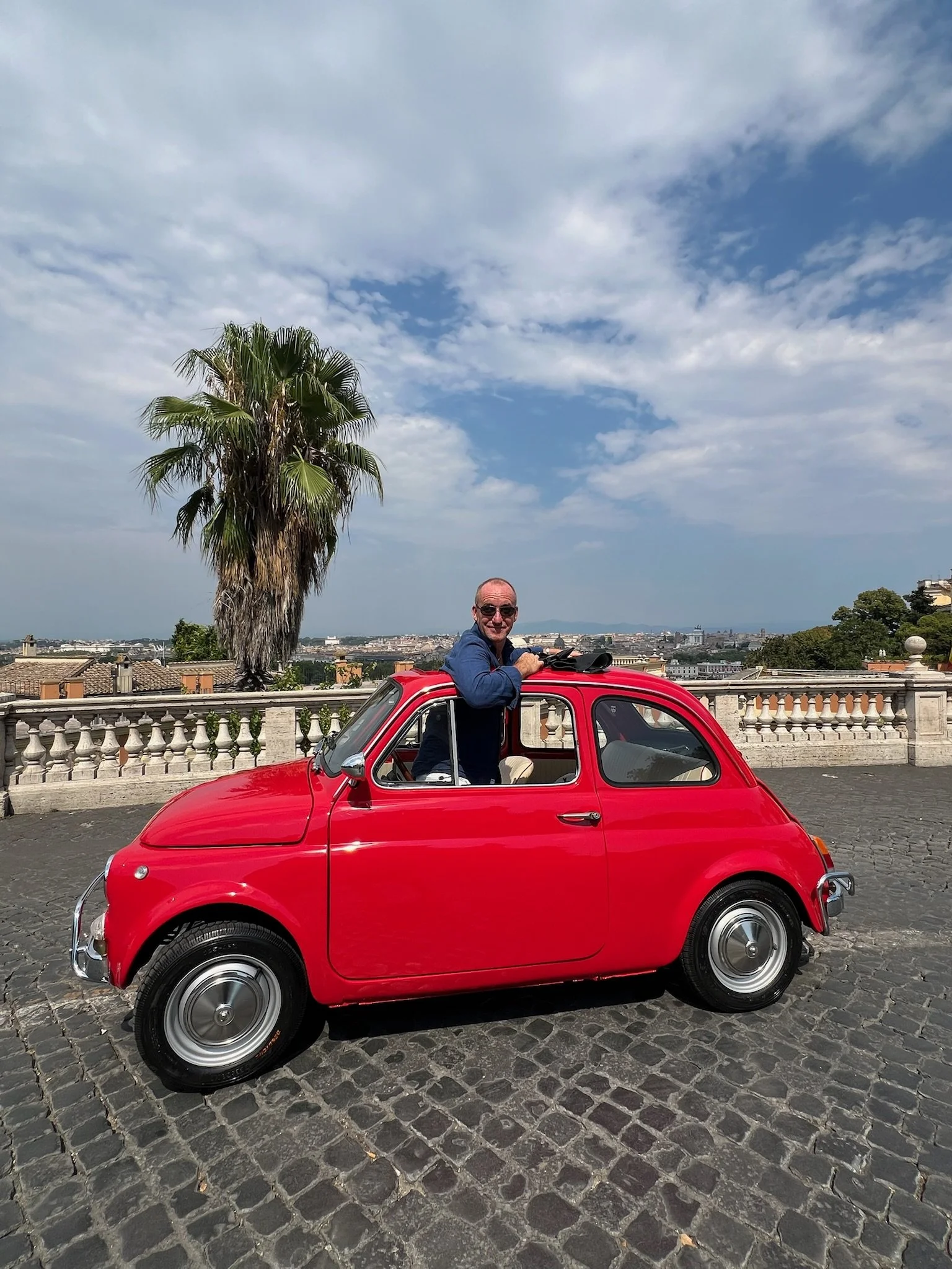 Uomo con occhiali seduto su una piccola auto rossa in una piazza con pavimentazione in pietra, alzato con il braccio, dietro un albero di palma e un parapetto con vista sulla città e il cielo con nuvole.