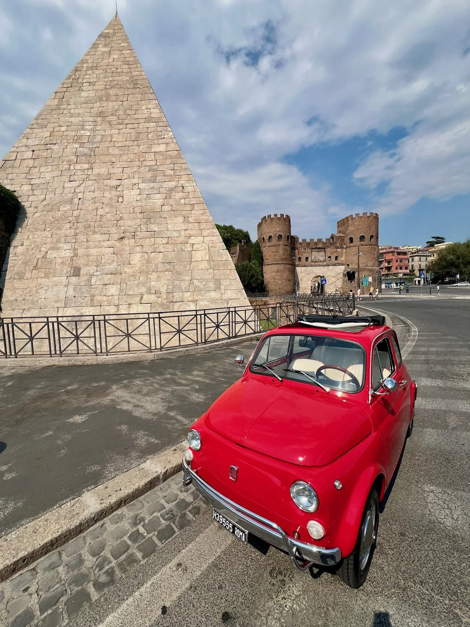 Una macchina rossa anni '60 parcheggiata su una strada con una piramide e un castello in pietra sullo sfondo, in un giorno con cielo parzialmente nuvoloso.