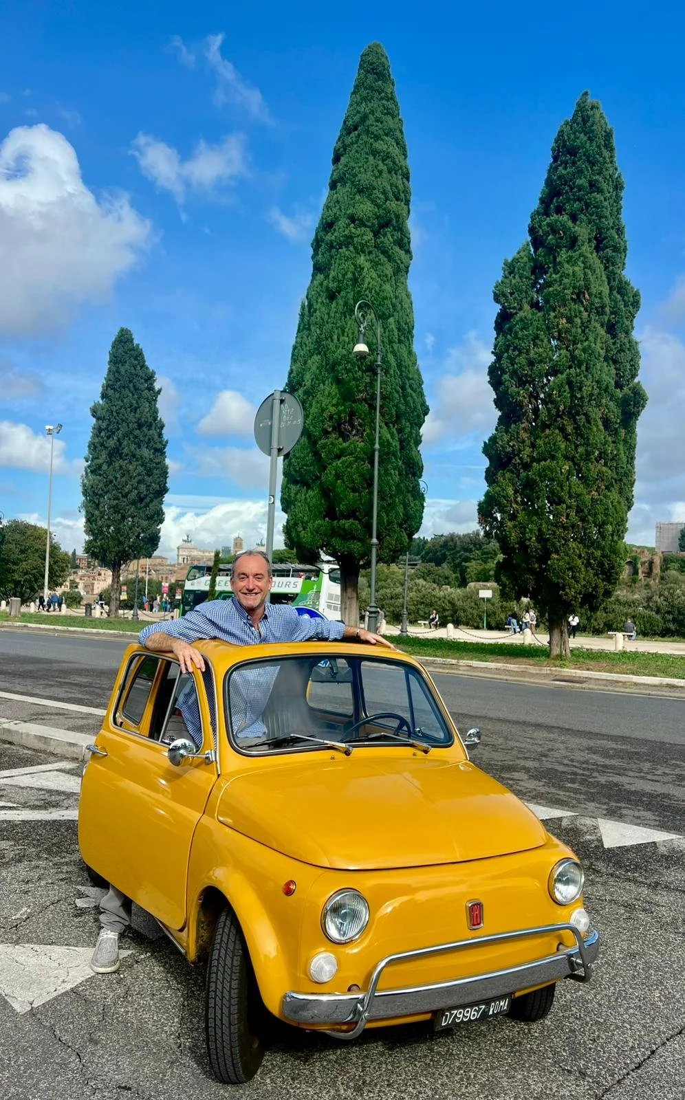Uomo con maglietta a maniche lunghe grigia e pantaloni chiari sorridente vicino a un'auto gialla vintage, con tre grandi alberi verdi sullo sfondo sotto un cielo blu con nuvole.
