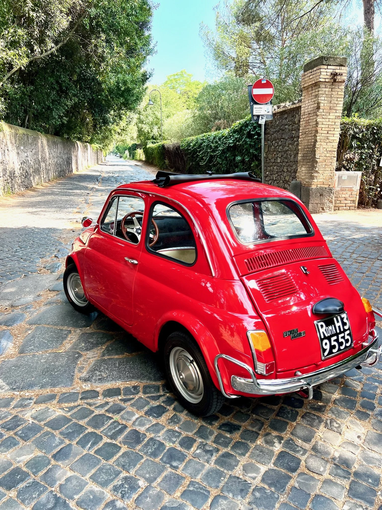 Auto d'epoca rosso parcheggiato su una strada in ciottoli, con segnali stradali e un muro di pietra in background.