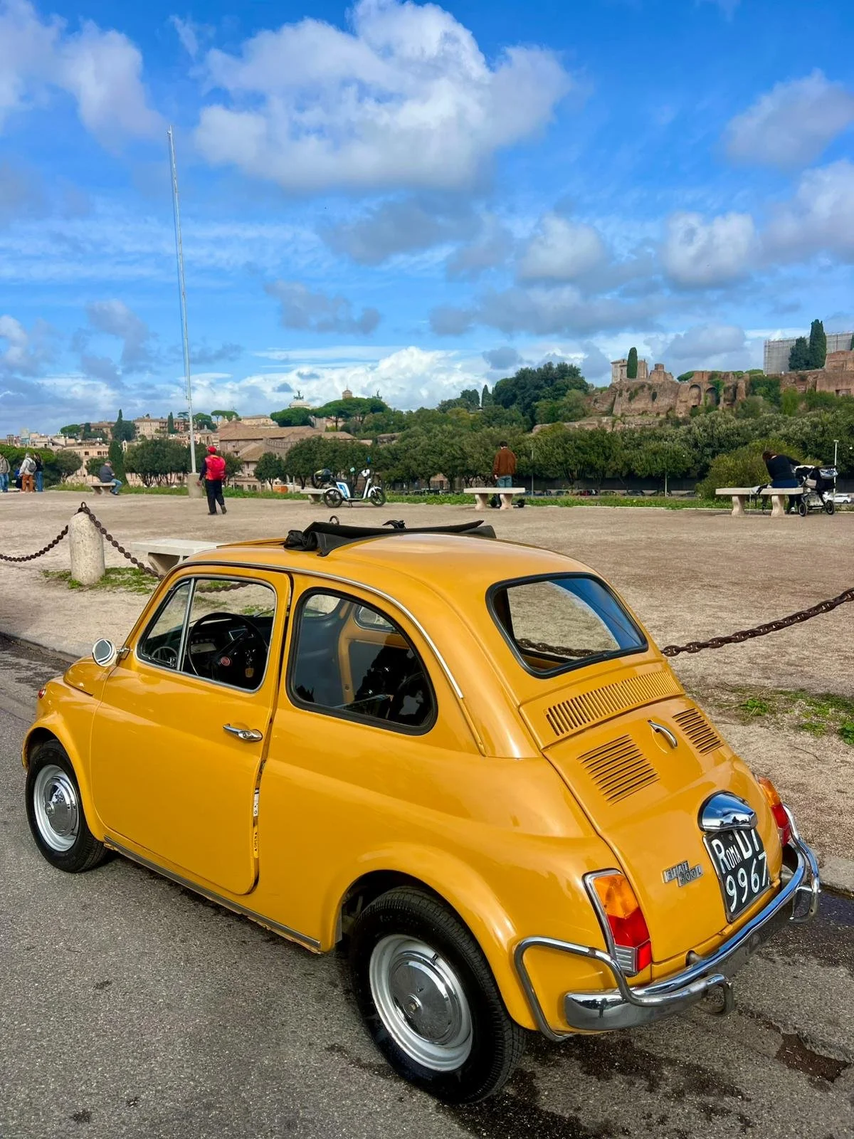 Auto d'epoca gialla parcheggiata in strada, con un paesaggio urbano e rovine storiche sullo sfondo, cielo sereno con nuvole.