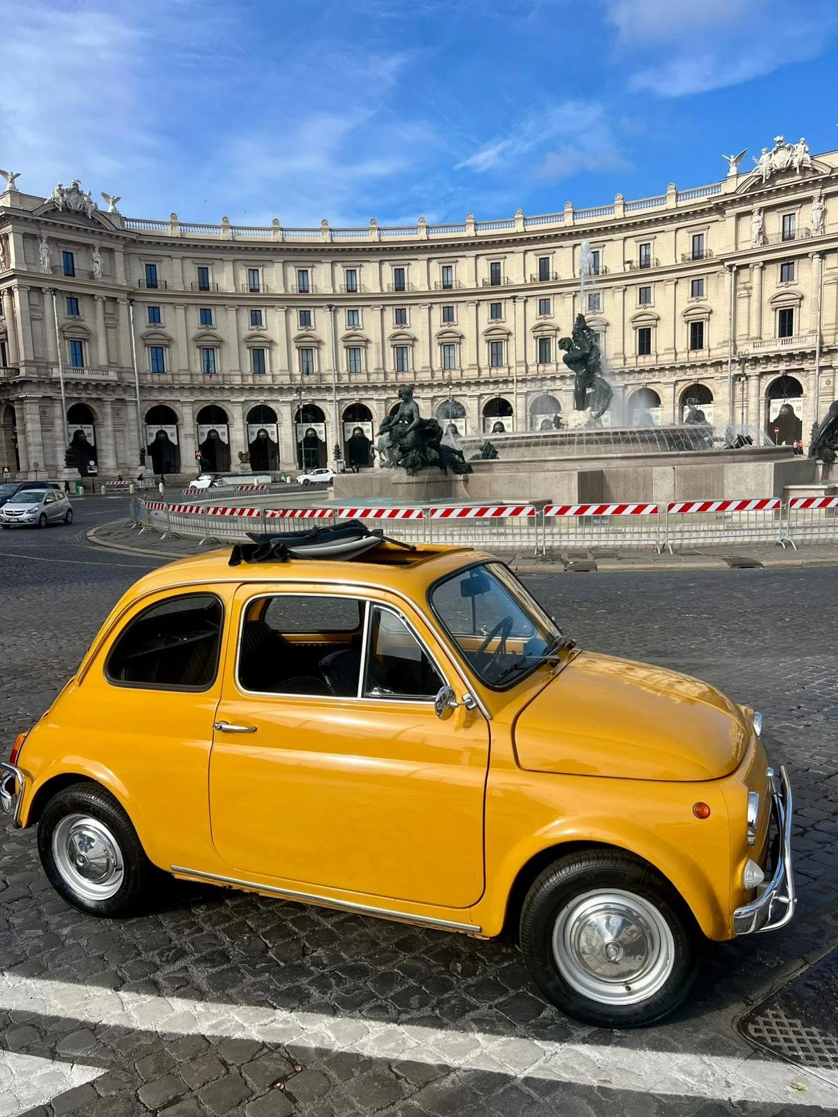 Una piccola auto gialla parcheggiata su un piazzale di pietra con un edificio storico e una fontana sulla sfondo di un cielo blu con poche nuvole.