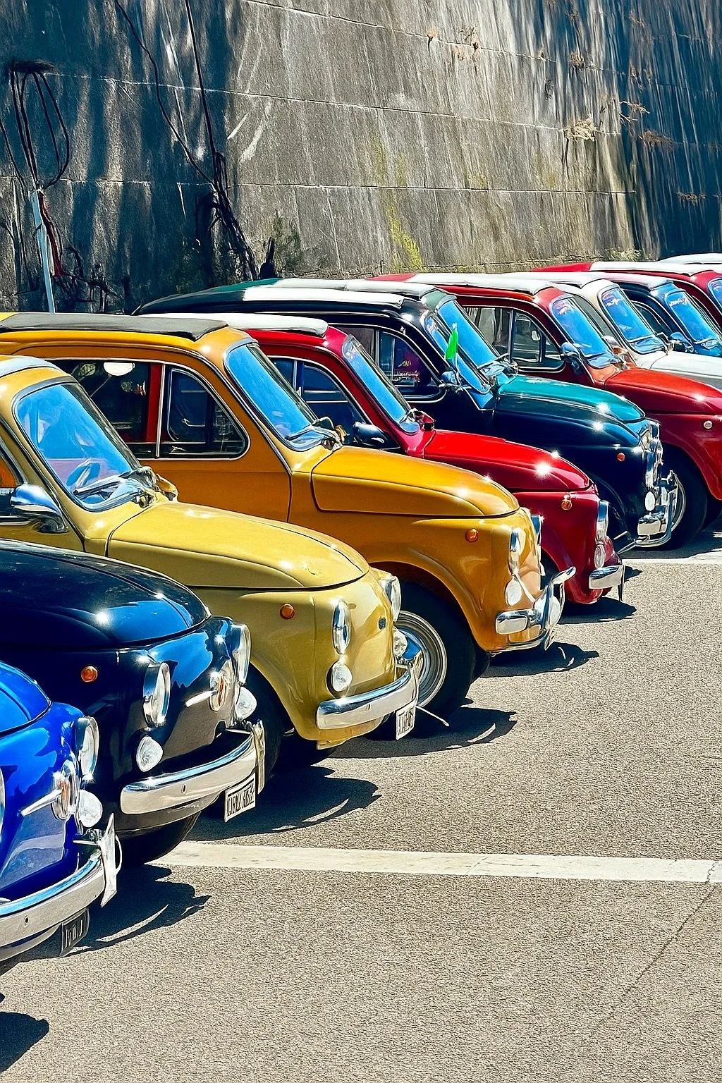 Una fila di auto colorate, principalmente gialle, rosse, blu e nere, parcheggiate lungo il bordo di una strada, con un muro di pietra sullo sfondo.