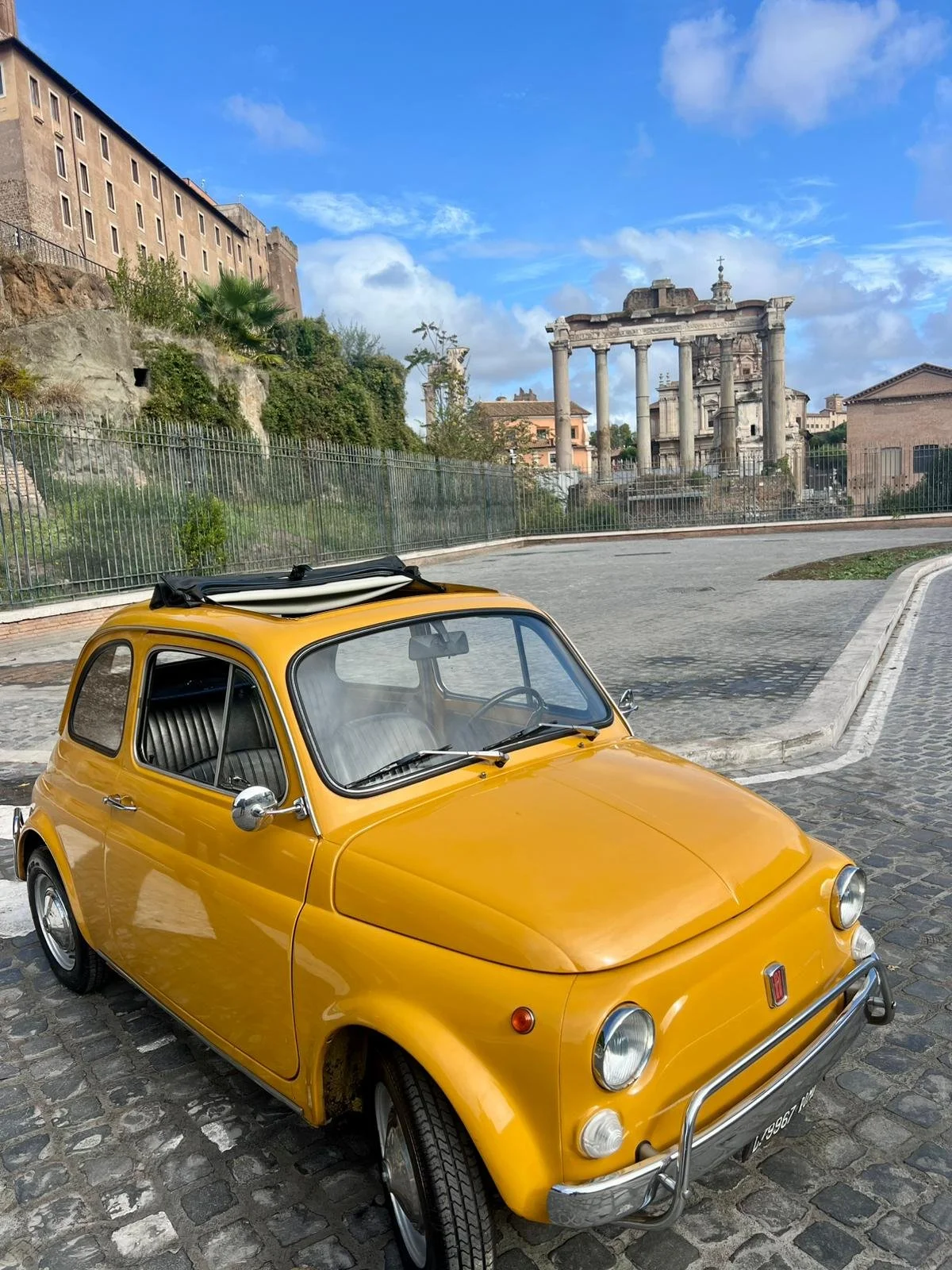 Auto giallo vintage parcheggiata su strada di ciottoli con rovine romane e edifici storici sullo sfondo, cielo sereno con nuvole sparse.